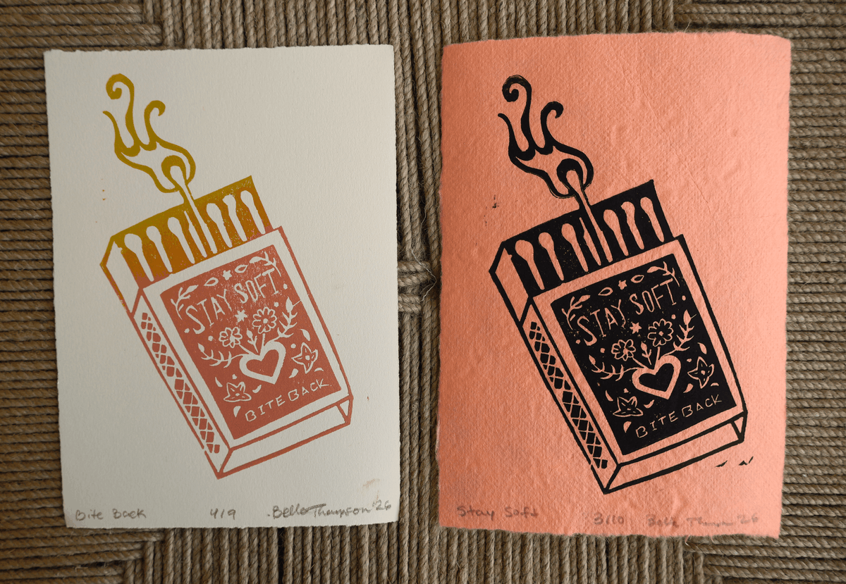 Two handmade art prints depicting a matchbox with a smoking match, side by side on a textured brown woven rug. The left print is on white paper with a muted red-orange matchbox and gold match head.