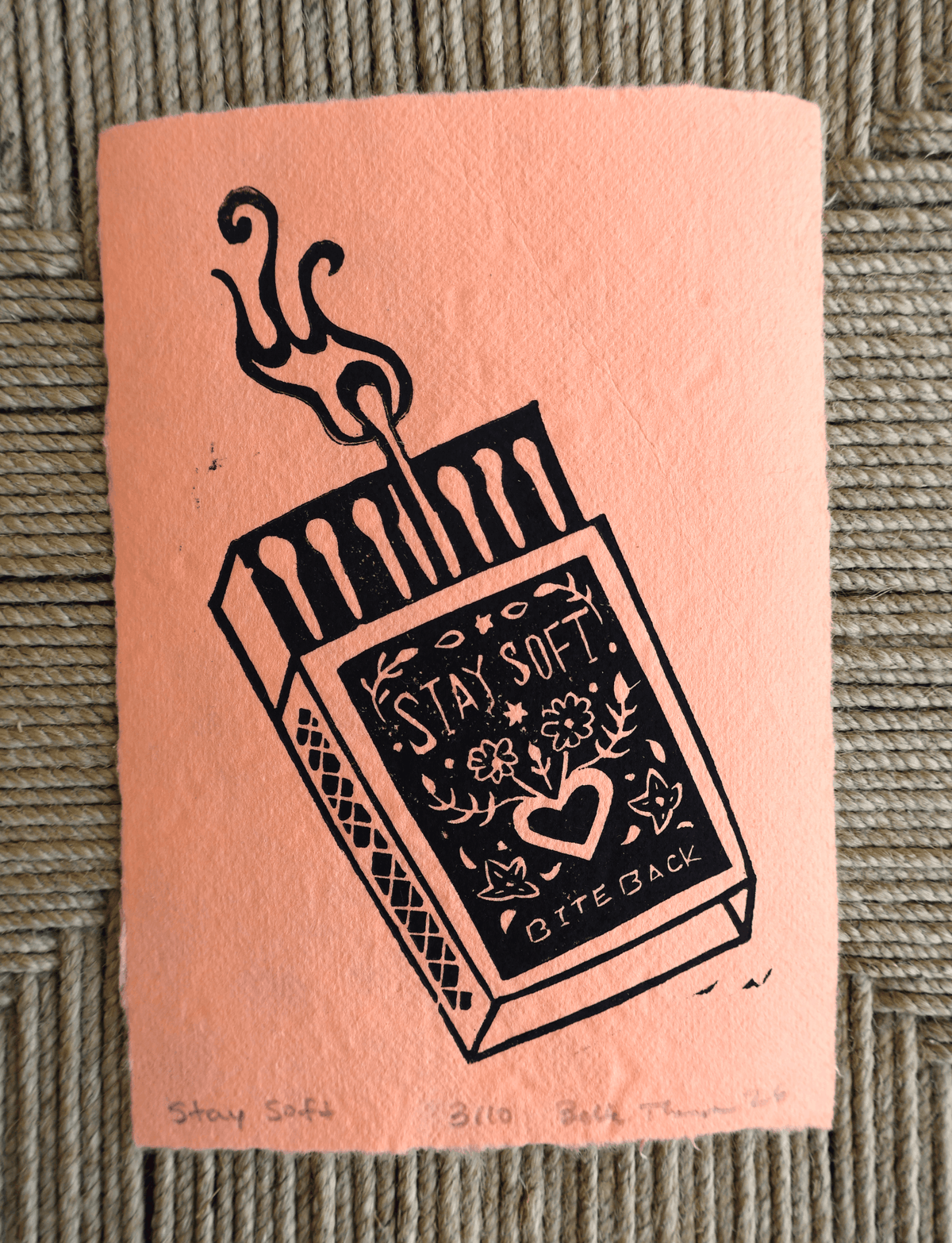 A square peach paper print or card featuring a black line-art illustration of an open matchbox with a hand reaching in and the words “STAY SOFT. BITE BACK.