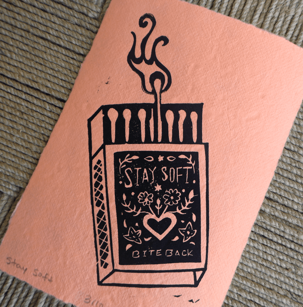 A square peach-colored art print or card featuring a black line drawing of an open matchbox with a flame and the words “STAY SOFT.” and “BITE BACK.