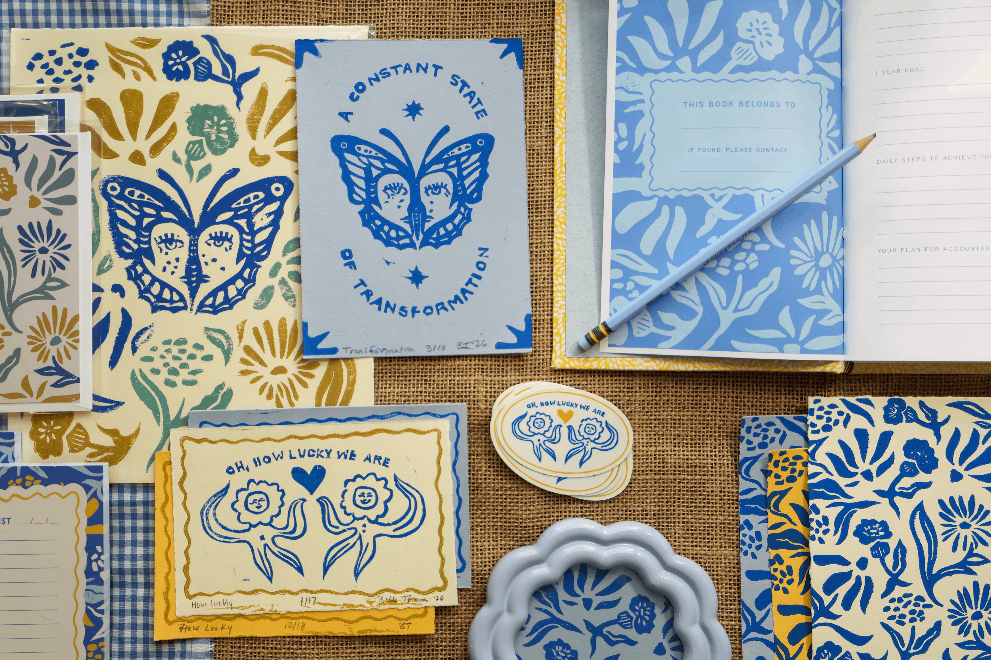 collection of metamorphosis collection stationery stickers self care journals grocery list stationery pas greeting cards handmade linocut prints and handbound books on a burlap and blue and white checkered background with saying of oh how lucky we are and a constant state of transformation with flower and butterfly imagery