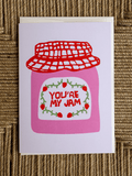 A square greeting card featuring a pink jam jar illustration with a red lid and the words 'YOU'RE MY JAM' in red, placed on a woven brown surface.