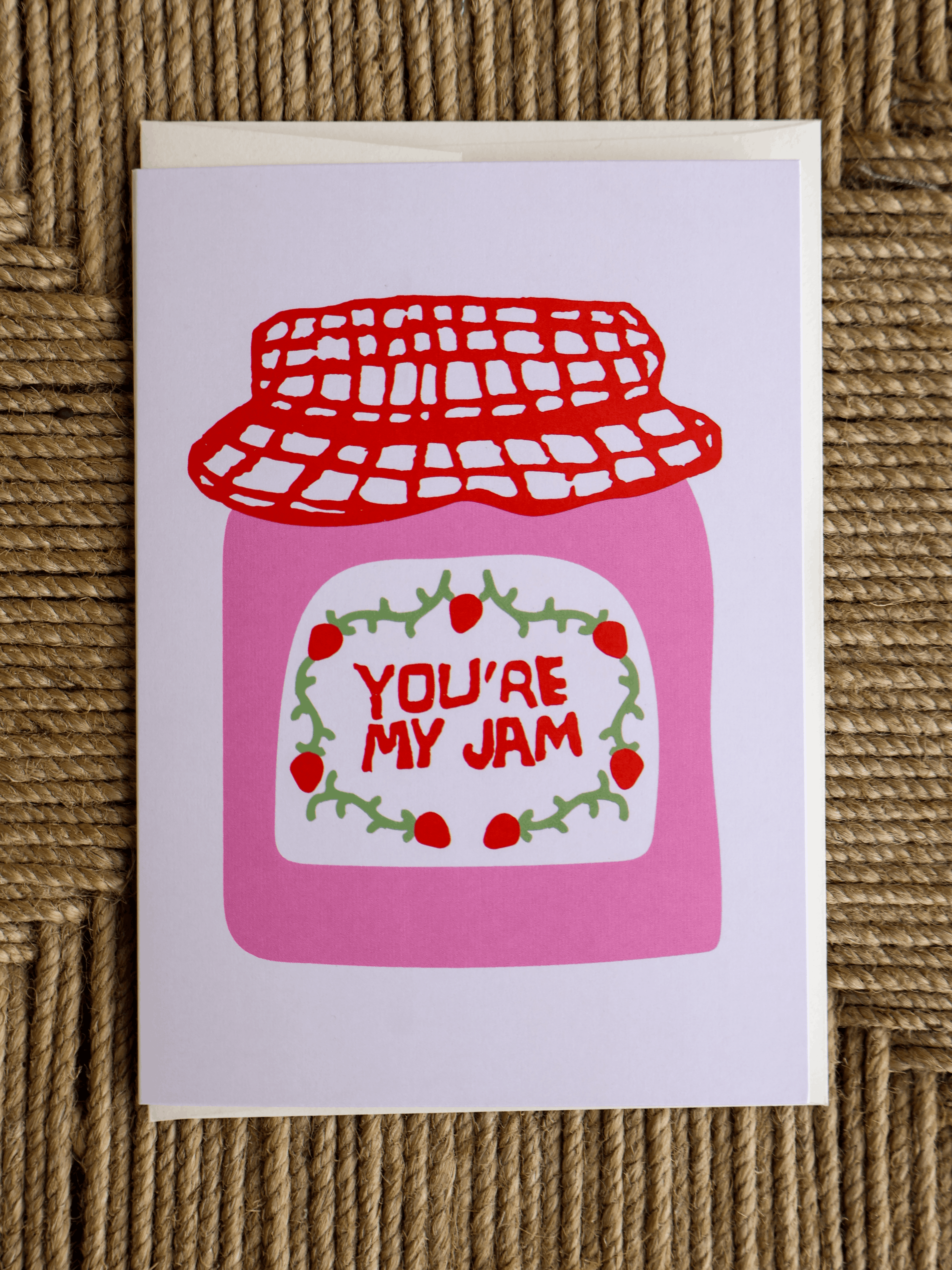 A square greeting card featuring a pink jam jar illustration with a red lid and the words 'YOU'RE MY JAM' in red, placed on a woven brown surface.