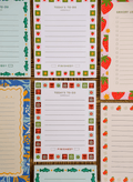 a colorful collection of handmade notepads arranged in a flat-lay on a woven natural background. The stationery features a variety of functional layouts, including to-do lists, grocery lists, and lined note pages, each framed with playful, nature-inspired illustrations such as strawberries, flowers, oranges, fish, and botanical motifs. The cohesive typography, vibrant yet earthy color palette, and charming decorative borders create a warm, artisan feel that highlights both the practicality and handcrafted. 