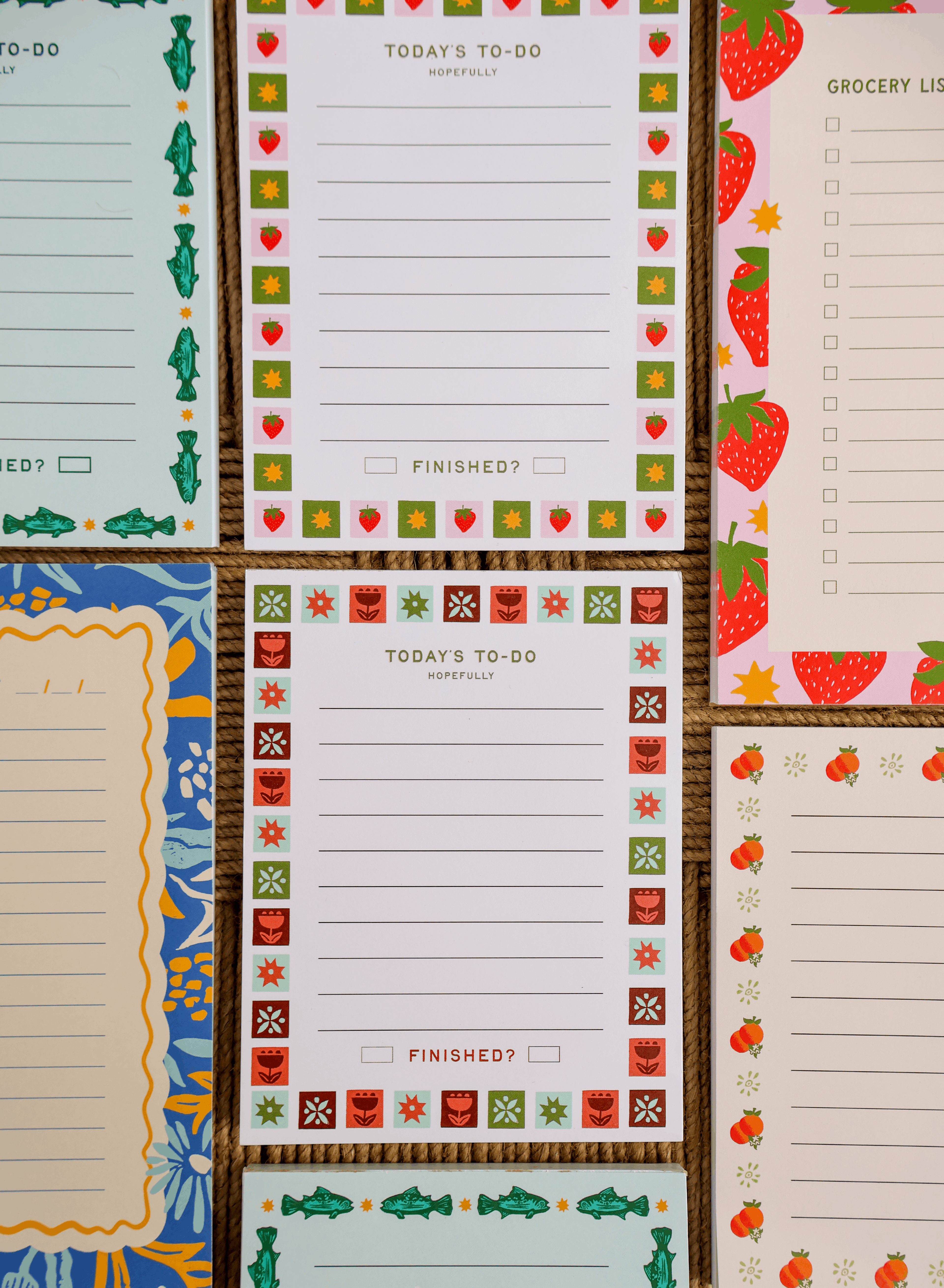 a colorful collection of handmade notepads arranged in a flat-lay on a woven natural background. The stationery features a variety of functional layouts, including to-do lists, grocery lists, and lined note pages, each framed with playful, nature-inspired illustrations such as strawberries, flowers, oranges, fish, and botanical motifs. The cohesive typography, vibrant yet earthy color palette, and charming decorative borders create a warm, artisan feel that highlights both the practicality and handcrafted. 