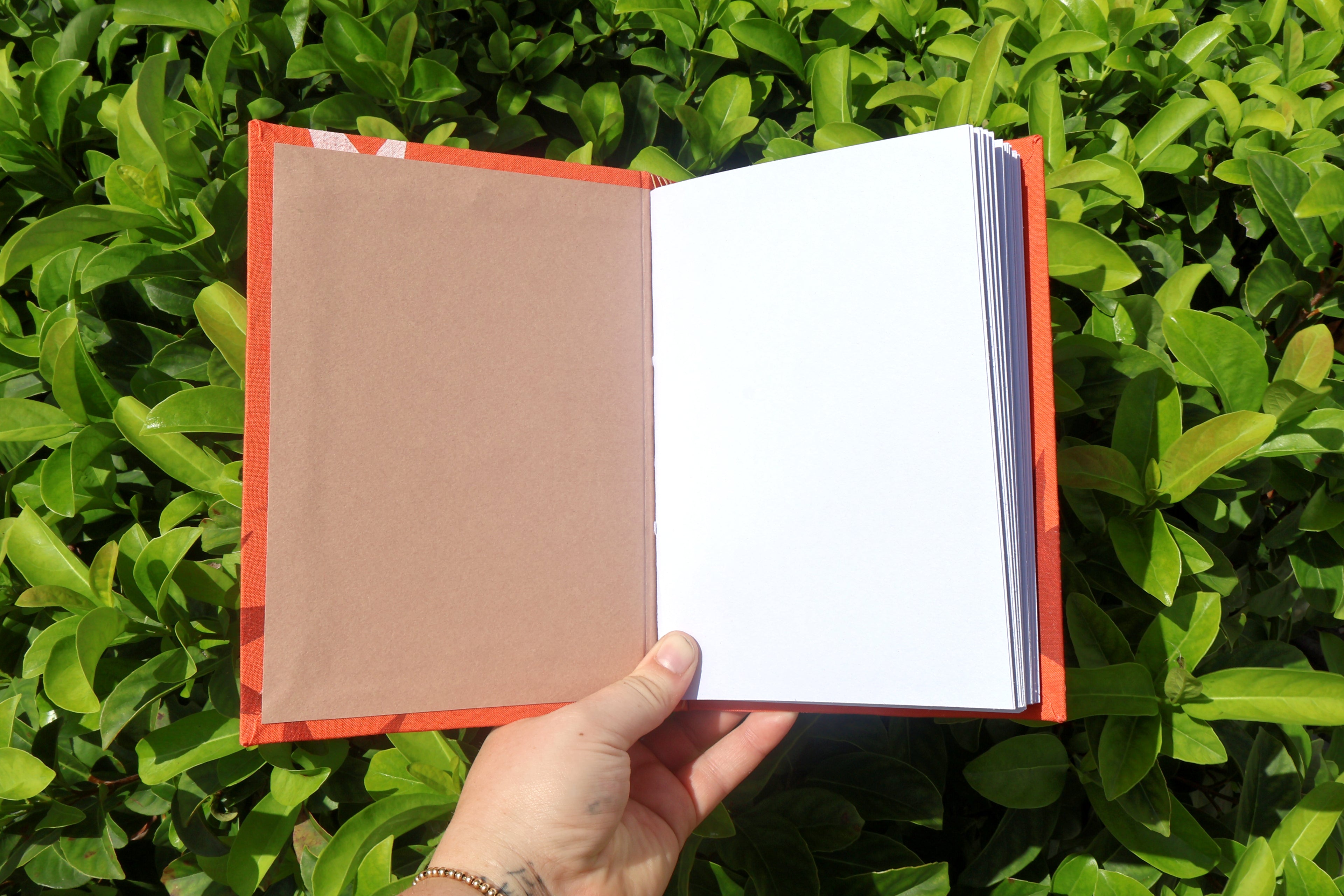 An open sketchbook with blank white watercolor paper pages on the right and a plain brown cover on the left, held by a hand against a background of green foliage.