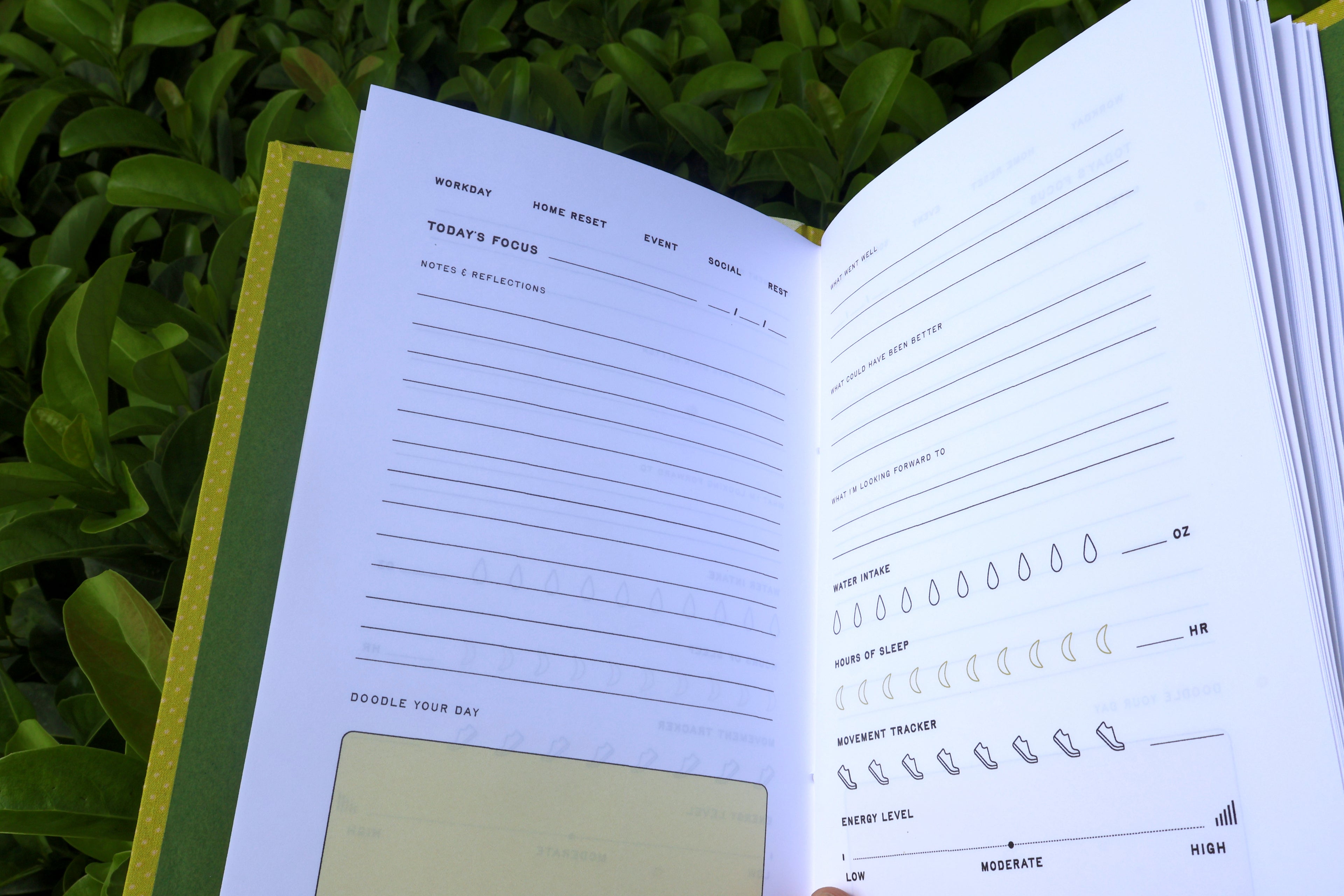An open daily journal or planner spread showing lined reflection pages with productivity trackers, photographed outdoors against green foliage. hand printed and bound self care reflections log