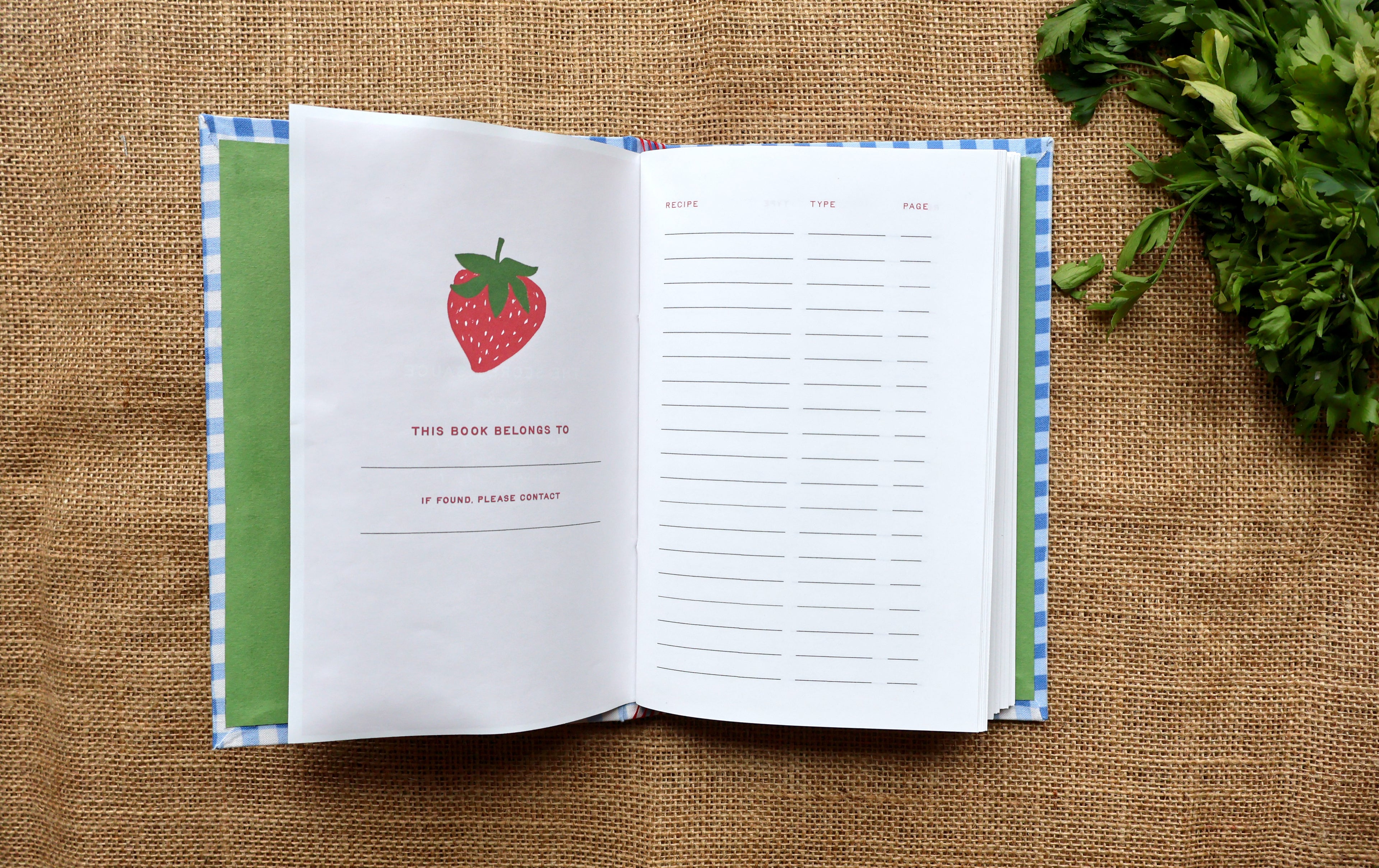 A handbound recipe log notebook with a blue and white gingham cover and a strawberry illustration on the front, shown open with recipe tracking pages inside and book claiming on a brown burlap background. 
