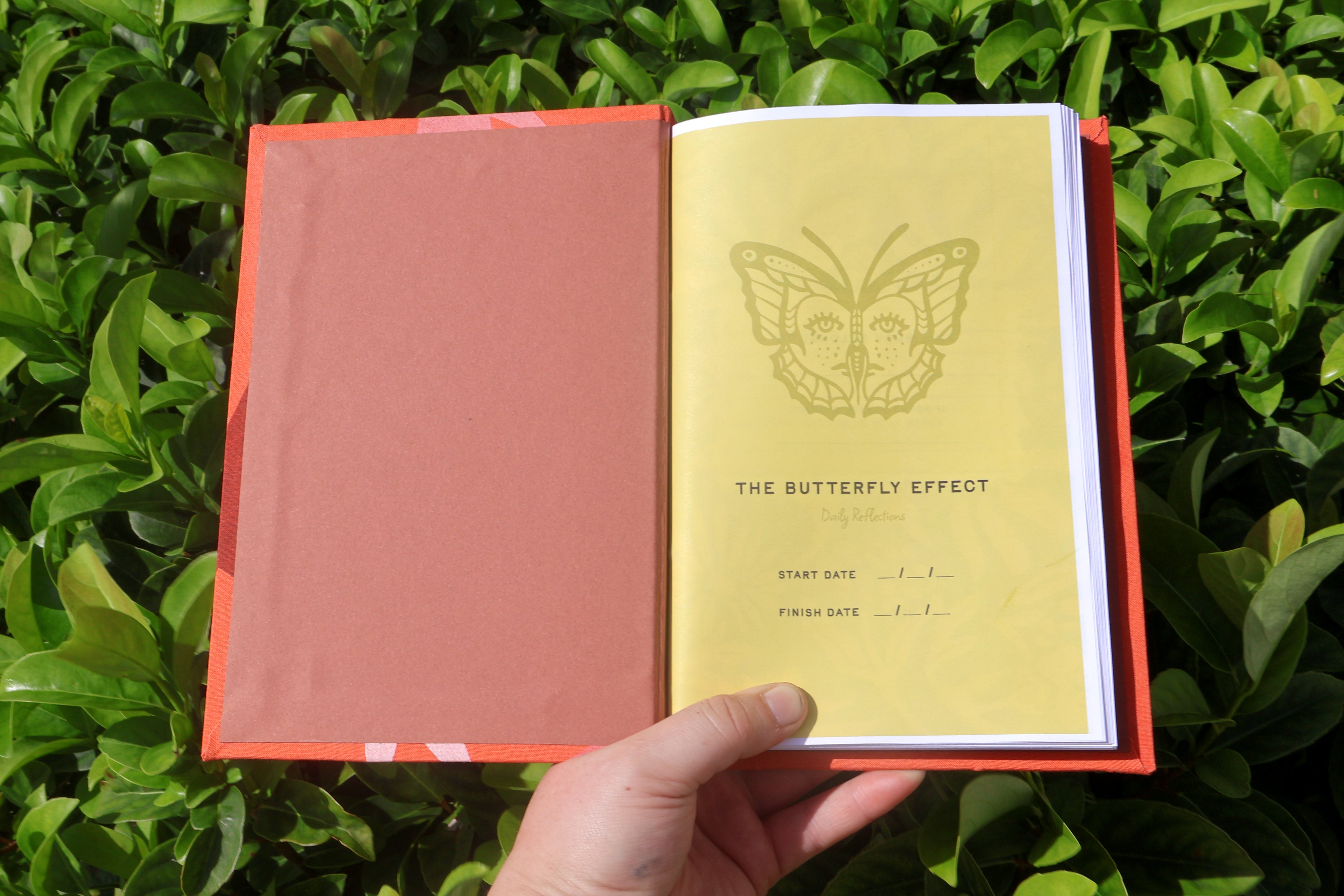 An open notebook or journal with a coral-red cover and a pale yellow inner page featuring a butterfly illustration and the title 'THE BUTTERFLY EFFECT.' A hand is holding it in front of dense green foliage. hand bound and linocut printed with more love studio
