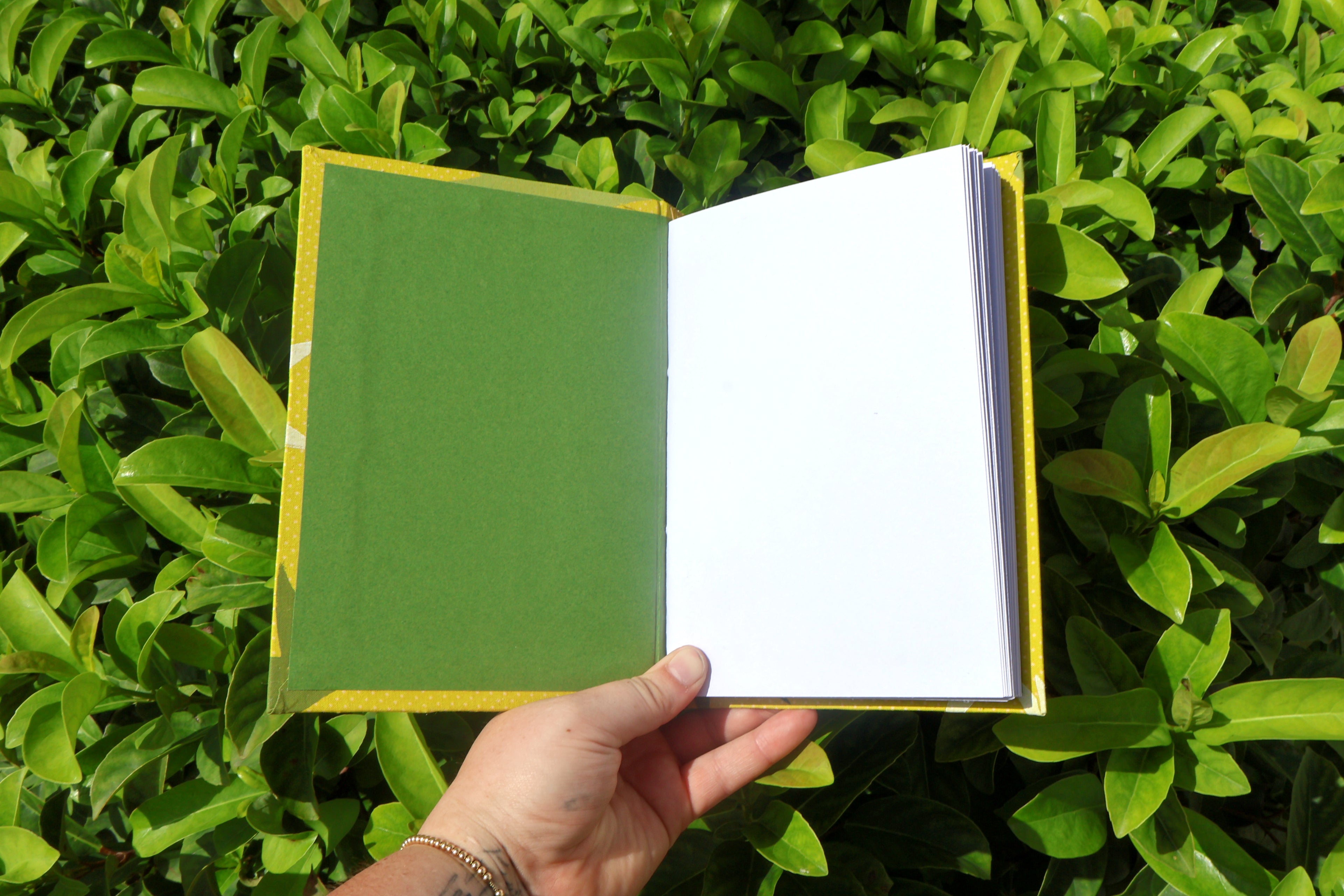 A hand holds an open, green, hand-bound and printed self-care reflection logs with white starburst shapes, photographed outdoors against dense green leaves. Hand bound and linocut printed with more love studio