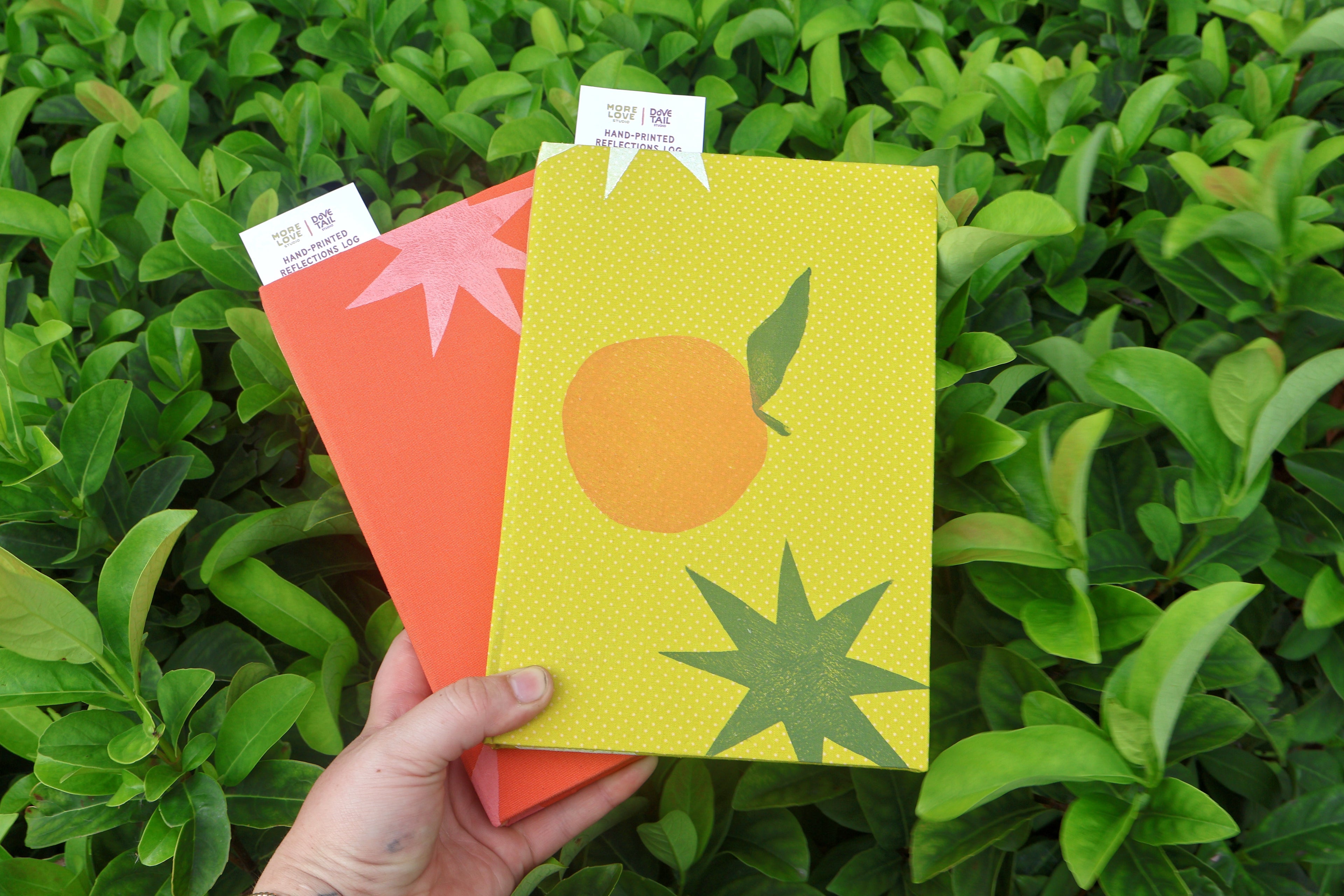 A hand holds two textured printed self care reflection logs in front of dense green foliage. The top card is bright yellow with a simple orange circle and green leaf, and the bottom card is coral-orange with a pink starburst. hand bound and printed with more love studio