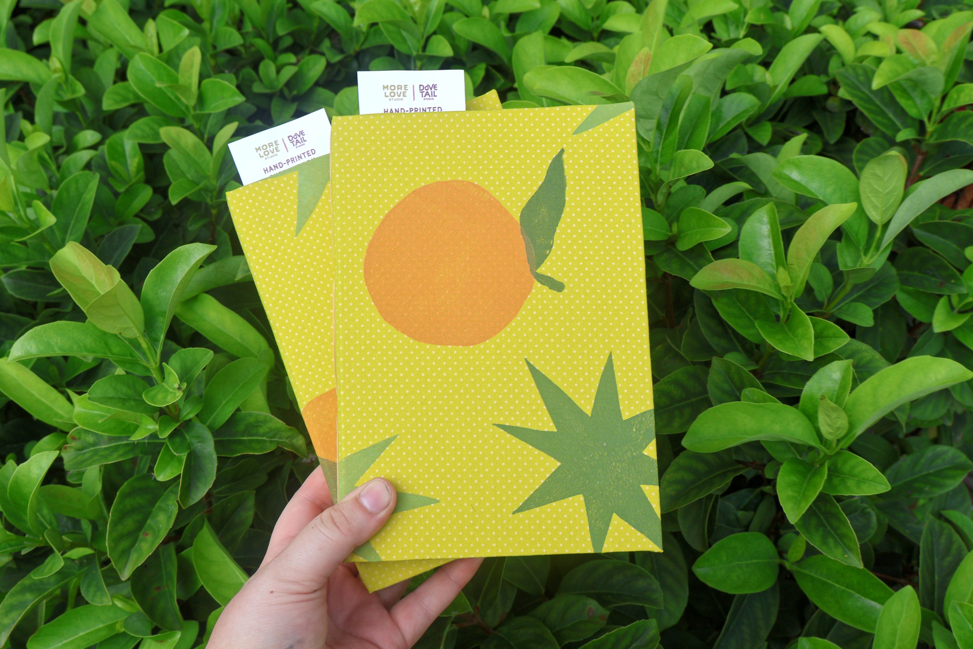A hand holds two hand printed and bound sketchbooks in front of dense green foliage. The front sketchbook is yellow with a white starburst and orange fruit, and the back journal is orange with a red starburst.