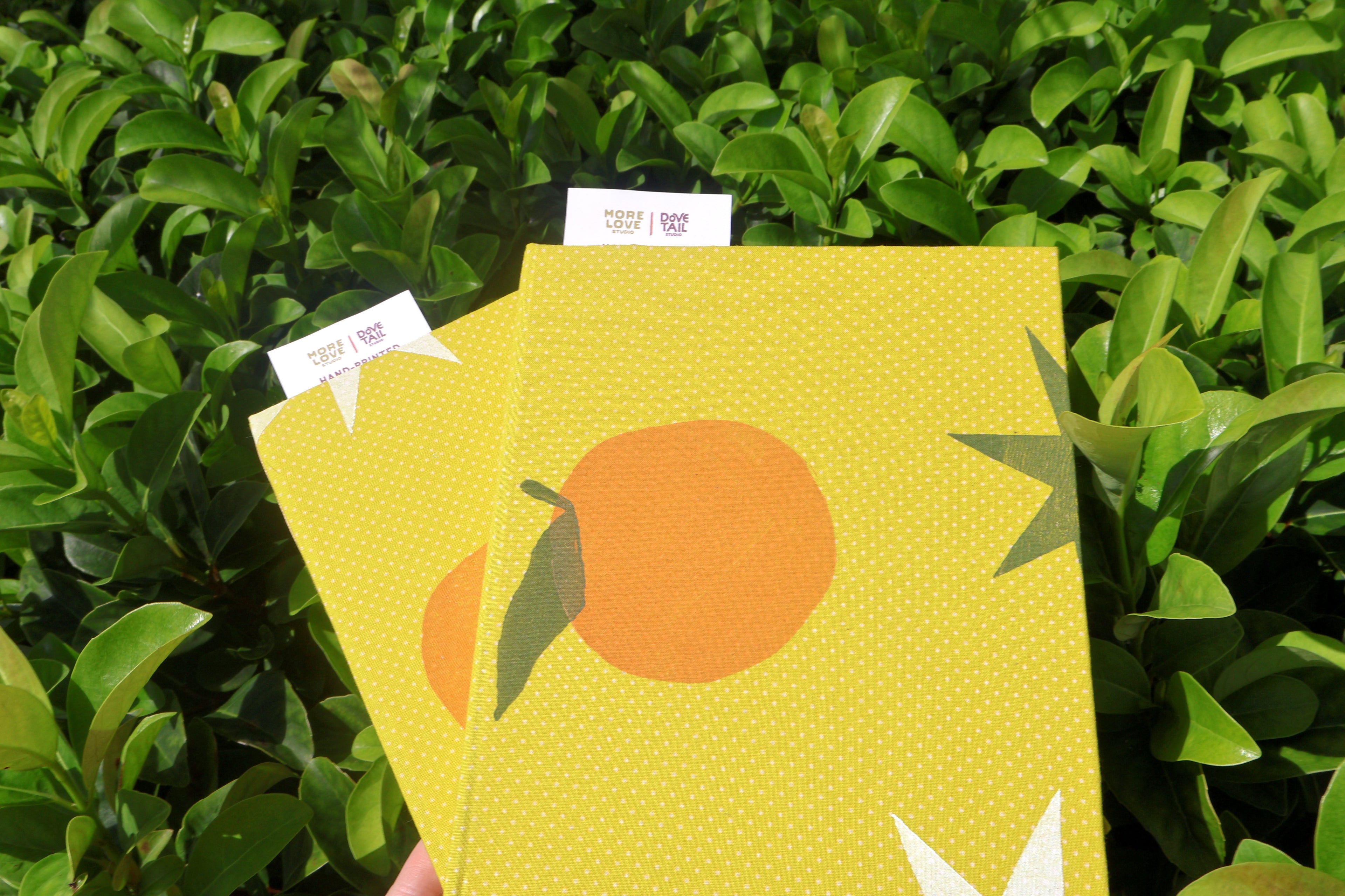 A yellow notebook with a dotted pattern and an orange graphic on the cover, held in front of a background of green leaves. The notebook has white tabs on the side.