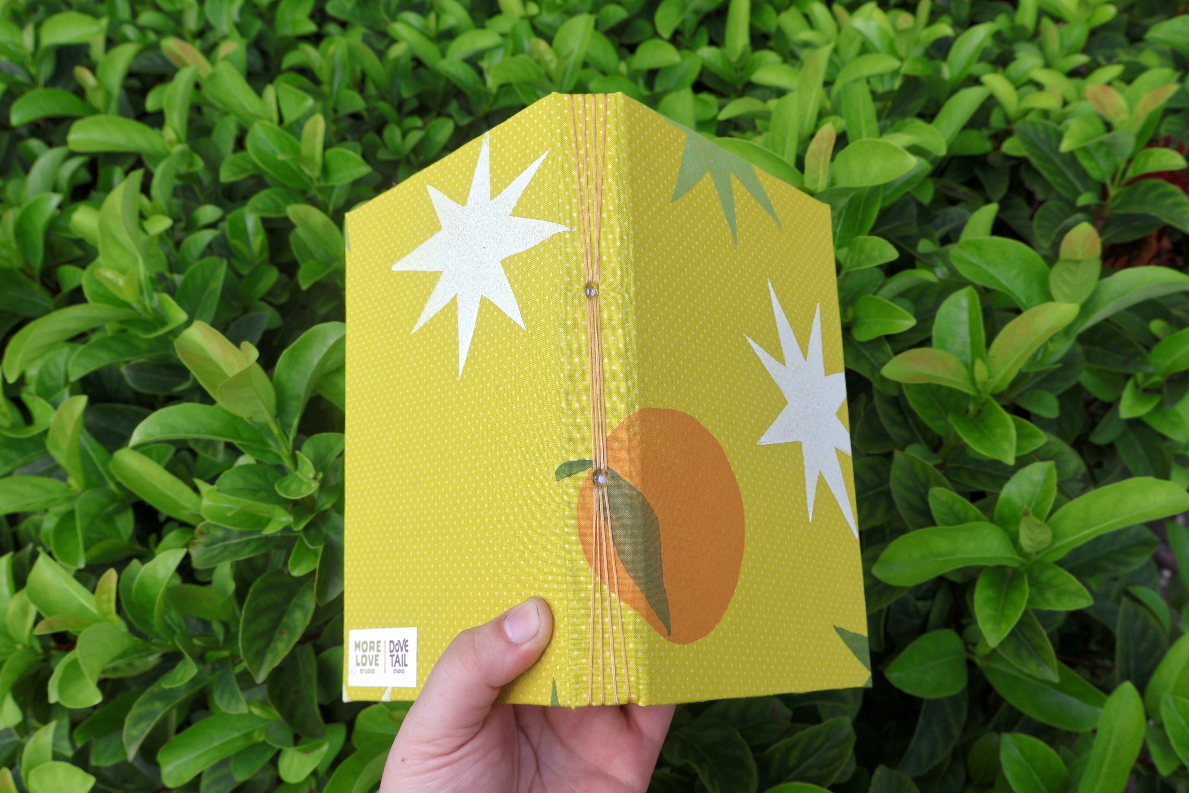A hand holds a small yellow sketchbook with a dotted texture, featuring white starburst graphics, a stylized orange, and green leaves. The background is a dense green hedge. Hand-bound and linocut printed with more love studio