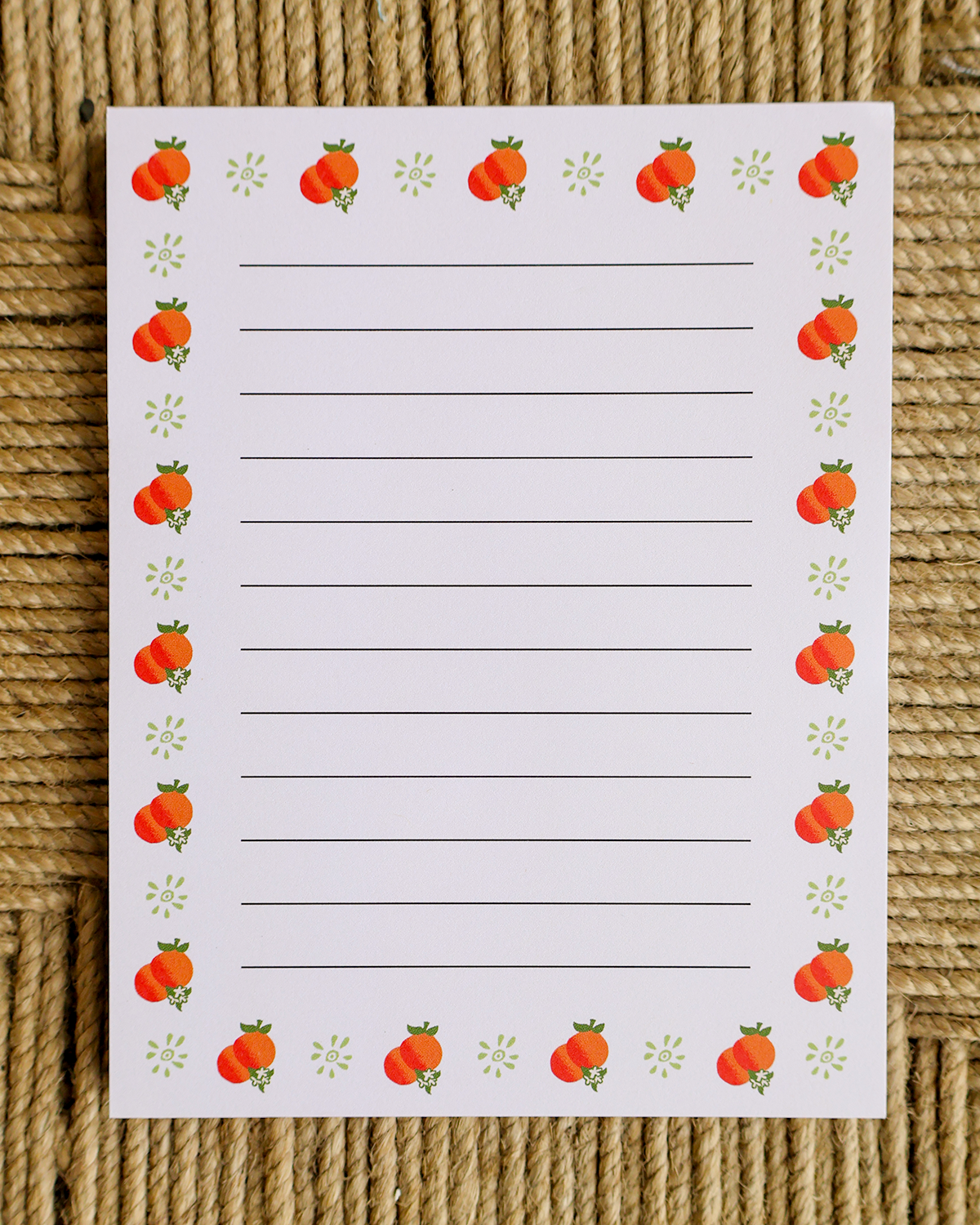 A lined writing pad with an orange and green border design, placed on a textured brown woven mat background.