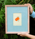 A framed art print held by hands against a background of green foliage. The print features a central cream-colored square with a colorful illustration of two oranges, surrounded by a light blue and white striped mat, all enclosed in a light pinkish-beige wooden frame.