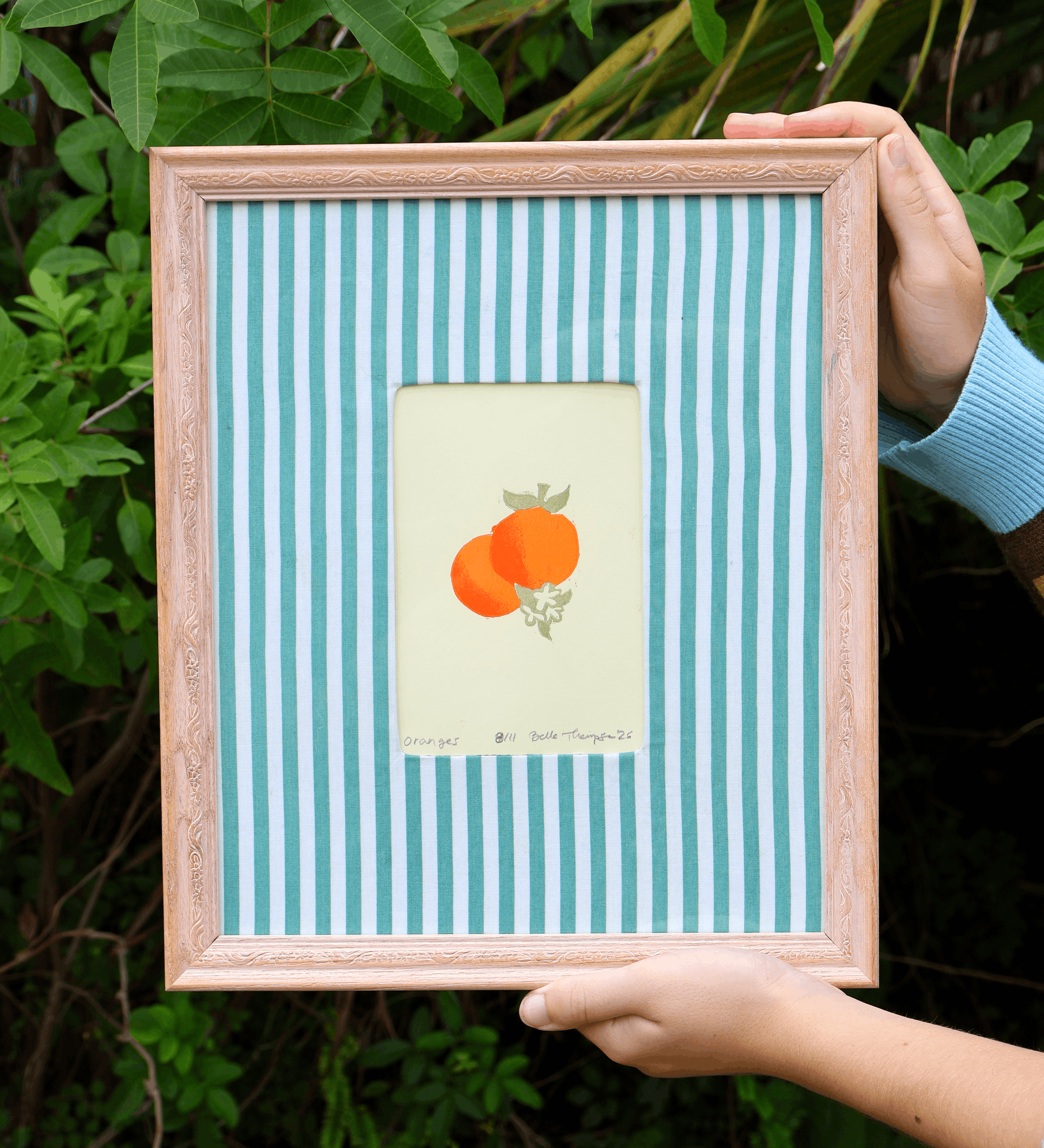A framed art print held by hands against a background of green foliage. The print features a central cream-colored square with a colorful illustration of two oranges, surrounded by a light blue and white striped mat, all enclosed in a light pinkish-beige wooden frame.