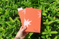 A hand holds two orange textured self care reflections logs decorated with orange and white starburst linocut prints, set against a background of green leaves. Hand printed linocut cover, hand bound self care reflections log