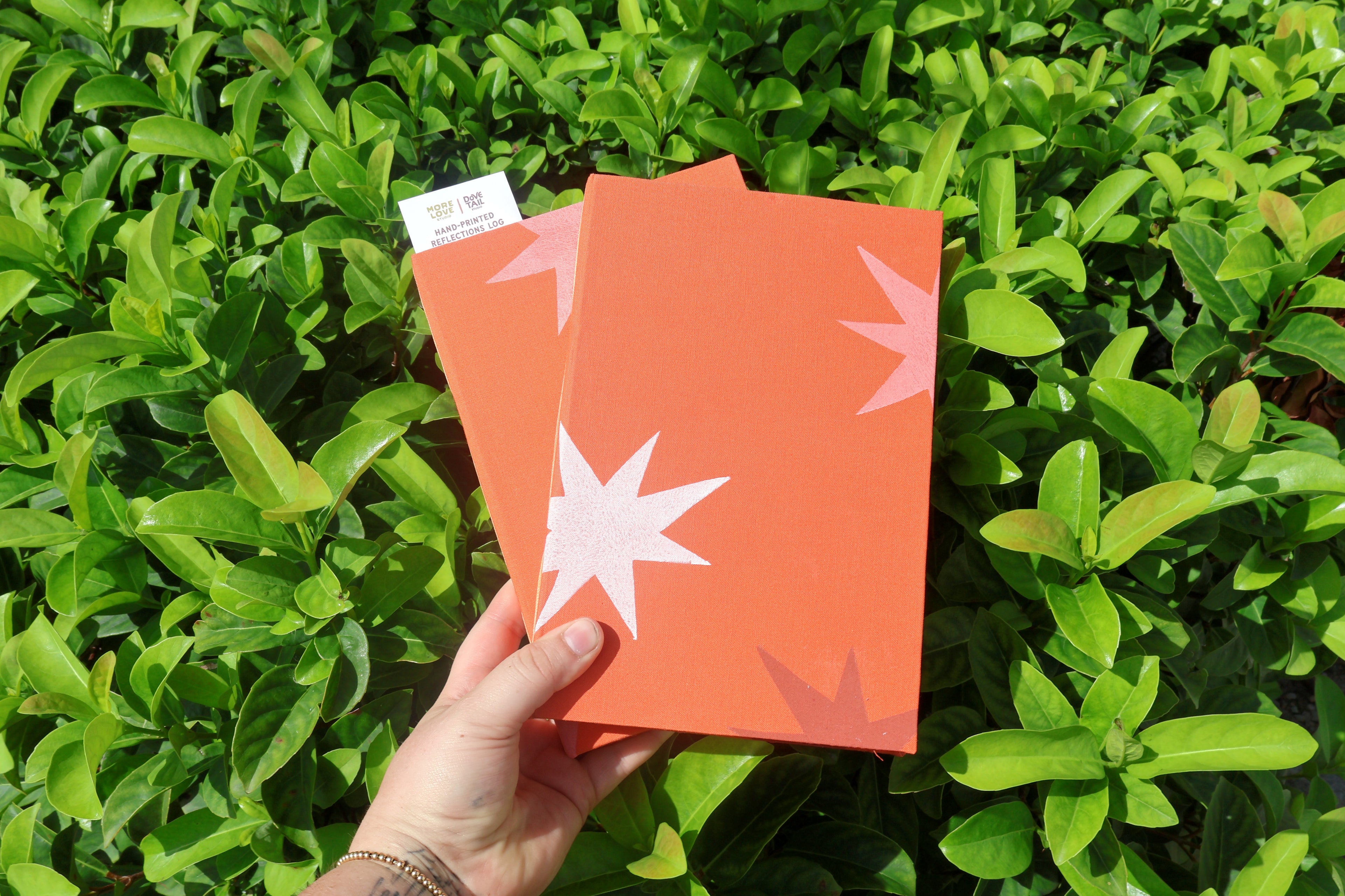 A hand holds two orange textured self care reflections logs decorated with orange and white starburst linocut prints, set against a background of green leaves. Hand printed linocut cover, hand bound self care reflections log
