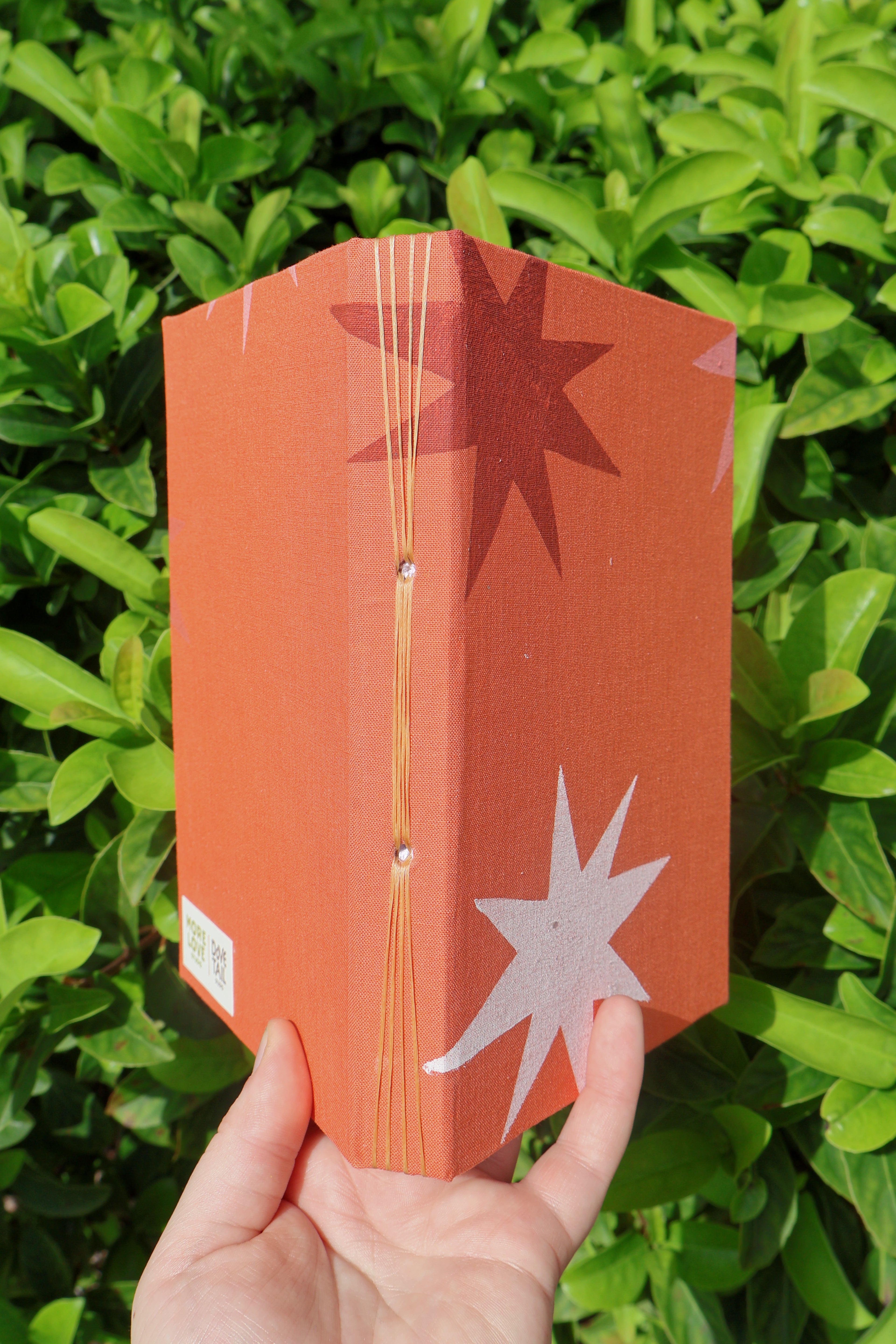 A hand holding a folded orange sketchbook with star cutouts, photographed outdoors against a background of green leaves. hand bound and linocut printed by more love studio