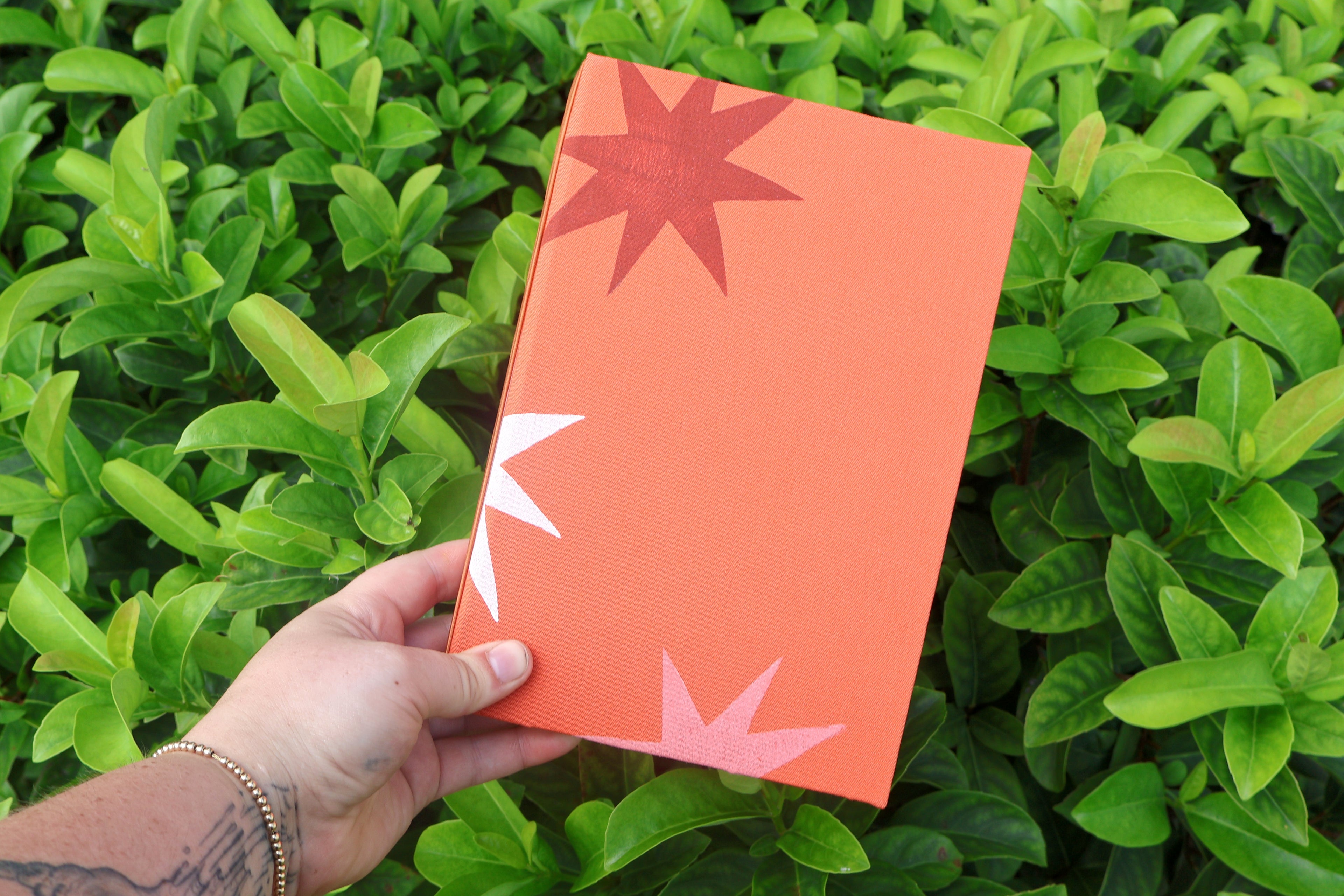 A hand holds a bright orange hardcover refillable journal with minimalist starburst cutout designs in red, white, and pink, set against a background of lush green leaves. hand bound and linocut printed with more love studio