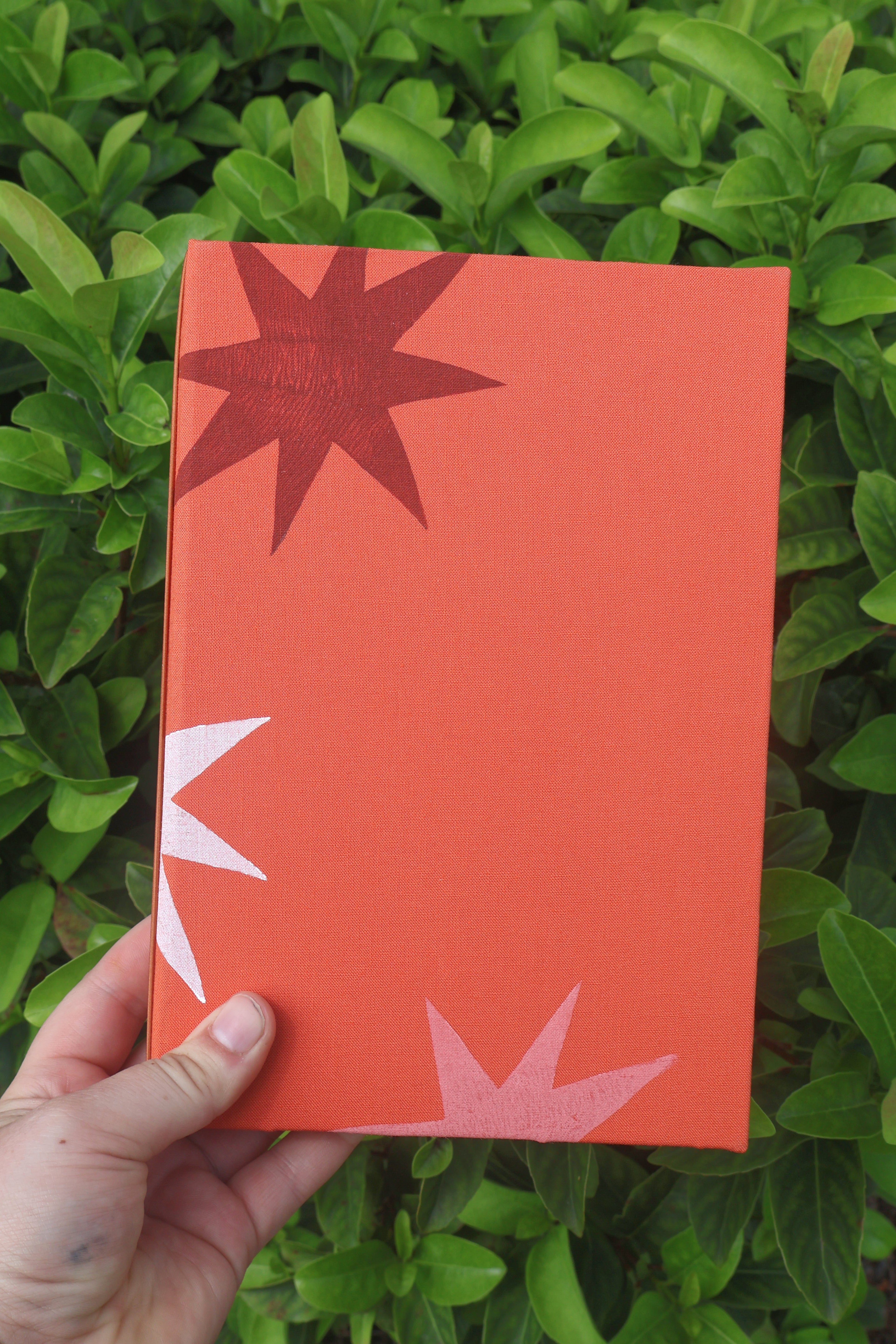 A hand holding a textured orange refillable journal cover with abstract starburst patterns in dark red, white, and light pink, set against a background of green leaves. hand bound and linocut printed with more love studio