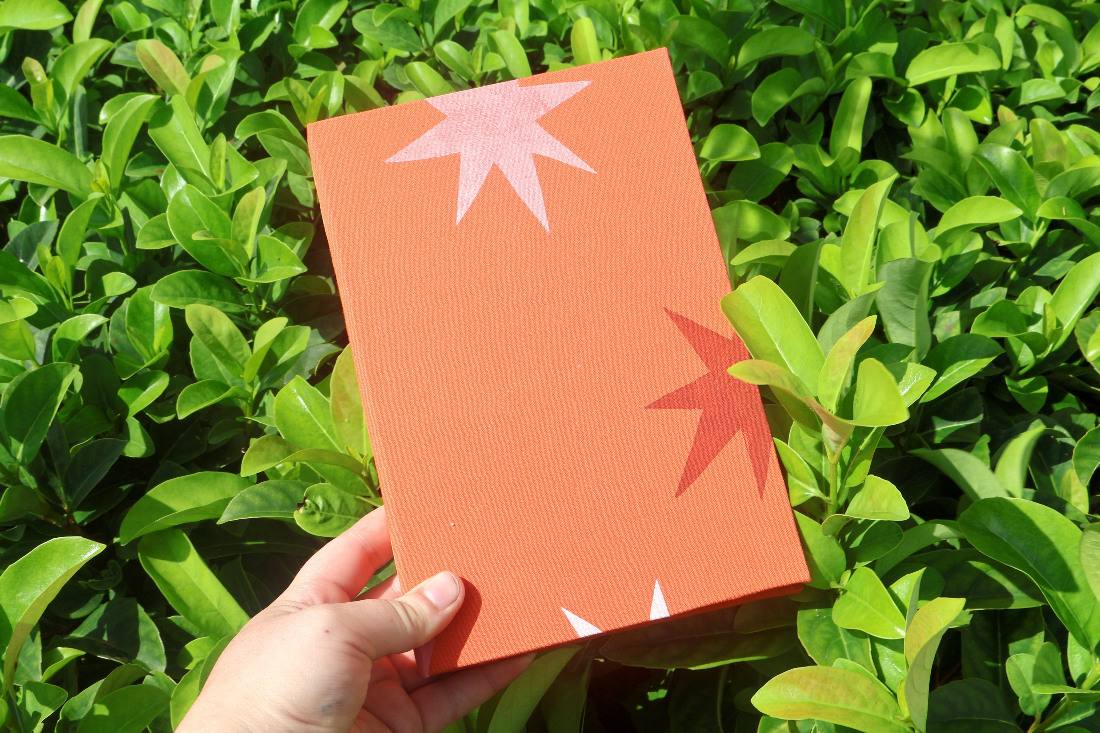 A hand holds an orange textured self care reflections logs decorated with orange and white starburst linocut prints, set against a background of green leaves. Hand printed linocut cover, hand bound self care reflections log