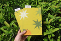 A hand holds two yellow patterned refillable journals in front of dense green foliage. The front journal has a white starburst, a dark green starburst, and a small orange shape, while the back journal has a white starburst and a green starburst. hand bound and linocut printed with more love studio