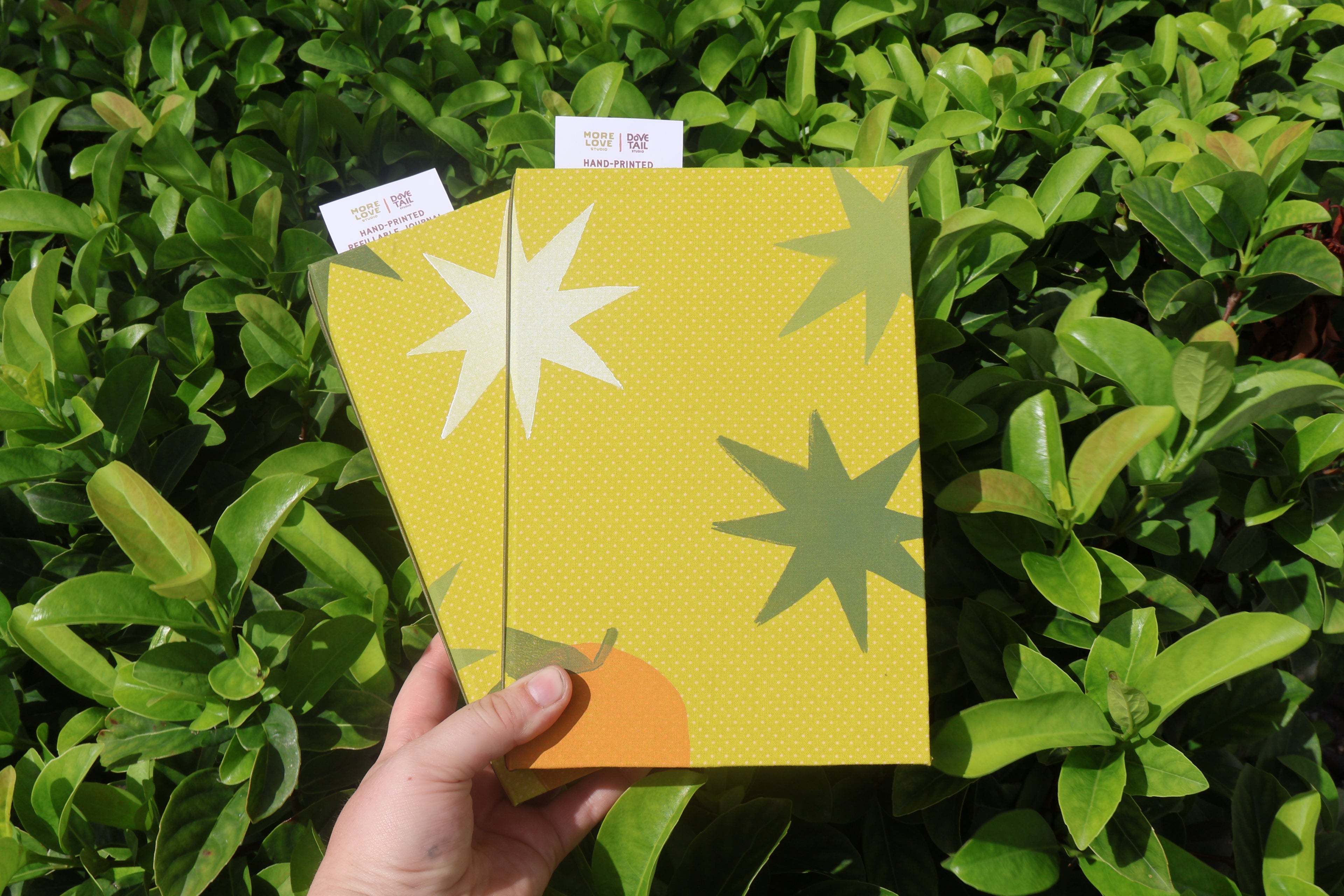 A hand holds two yellow patterned refillable journals in front of dense green foliage. The front journal has a white starburst, a dark green starburst, and a small orange shape, while the back journal has a white starburst and a green starburst. hand bound and linocut printed with more love studio