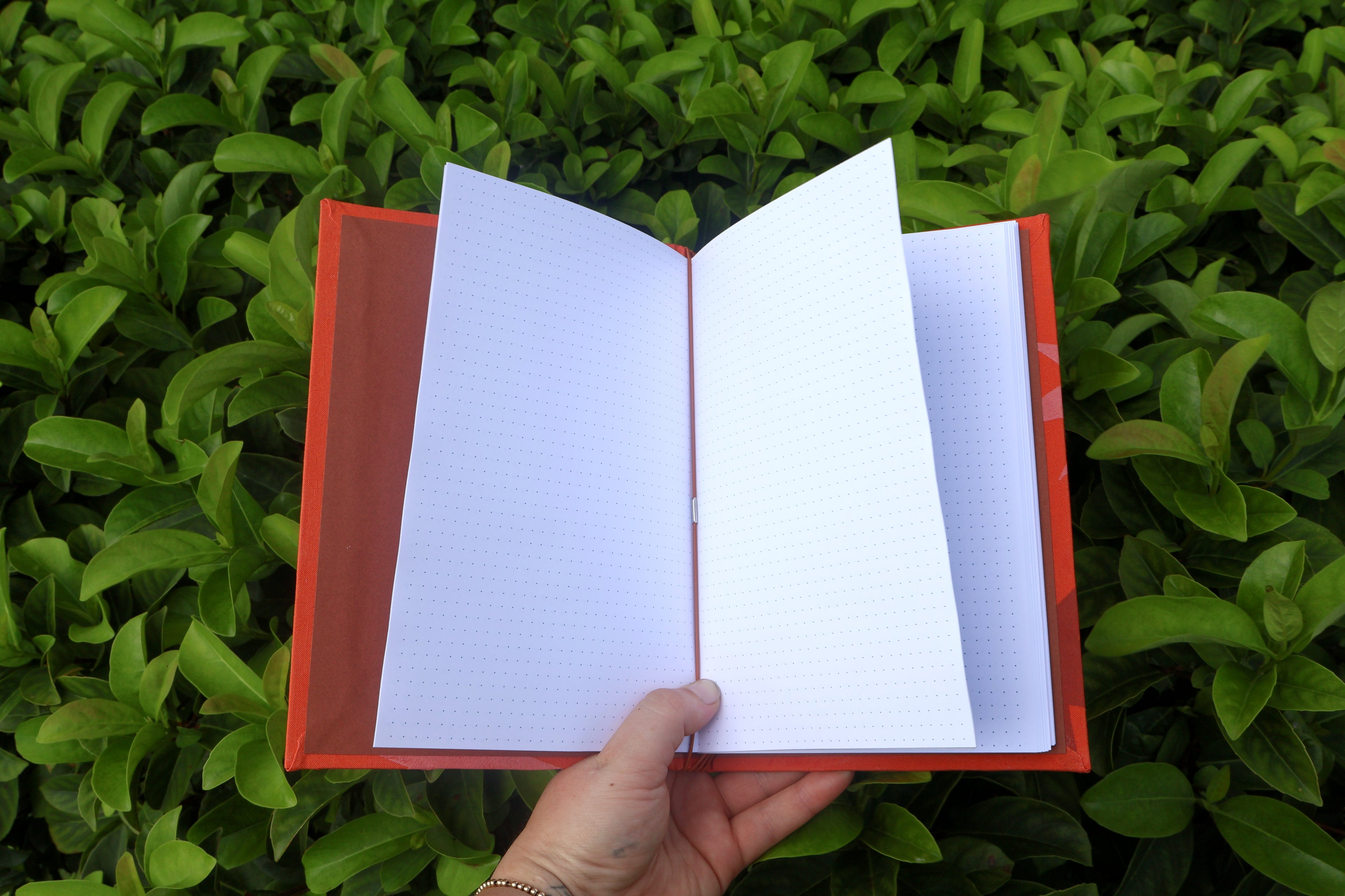 An open orange starburst refillable journal with white grid pages held by a hand against a background of green foliage. hand bound and linocut printed with more love studio