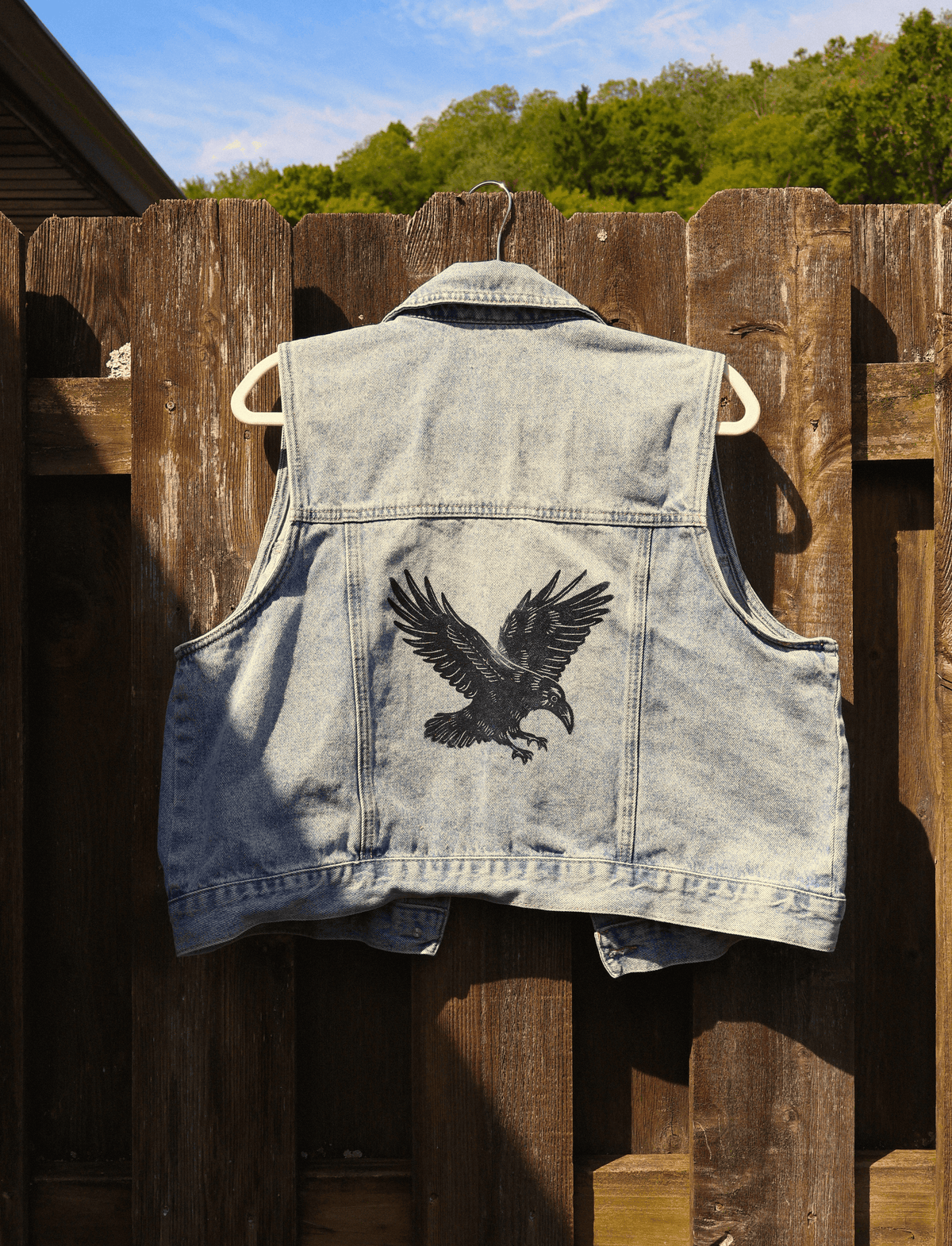 A light blue denim vest hanging on a wooden fence outdoors, featuring a large black eagle graphic printed on the back.