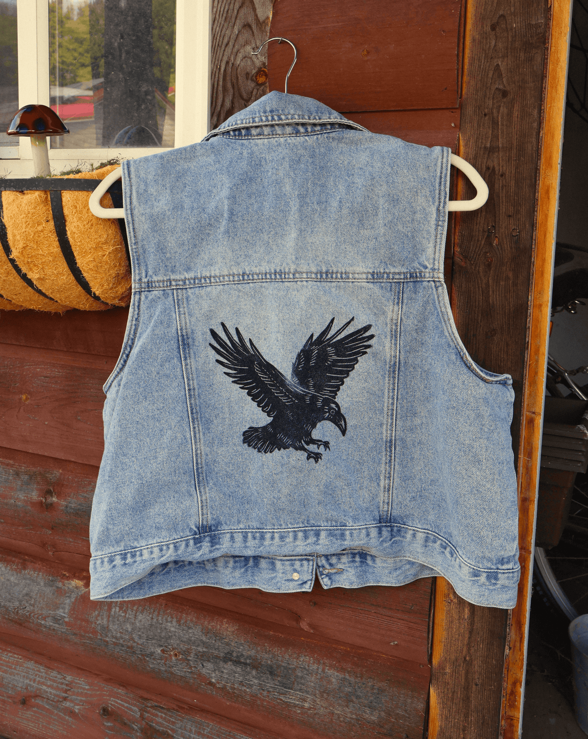 A light blue denim vest hanging on a white hanger against a rustic wooden door, featuring a large black embroidered raven design on the back.