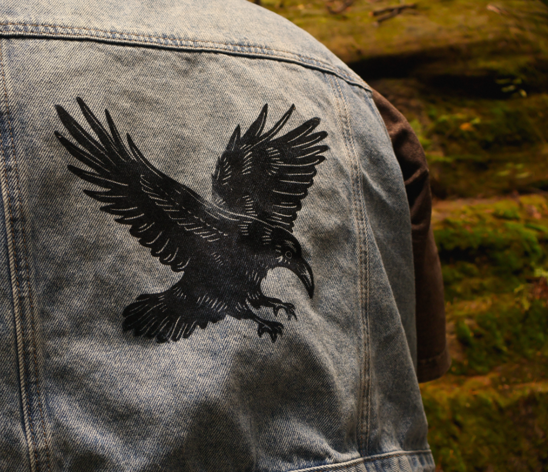 Close-up of the back of a light blue denim jacket featuring a large black embroidered raven design, worn outdoors with a blurred mossy rock background.