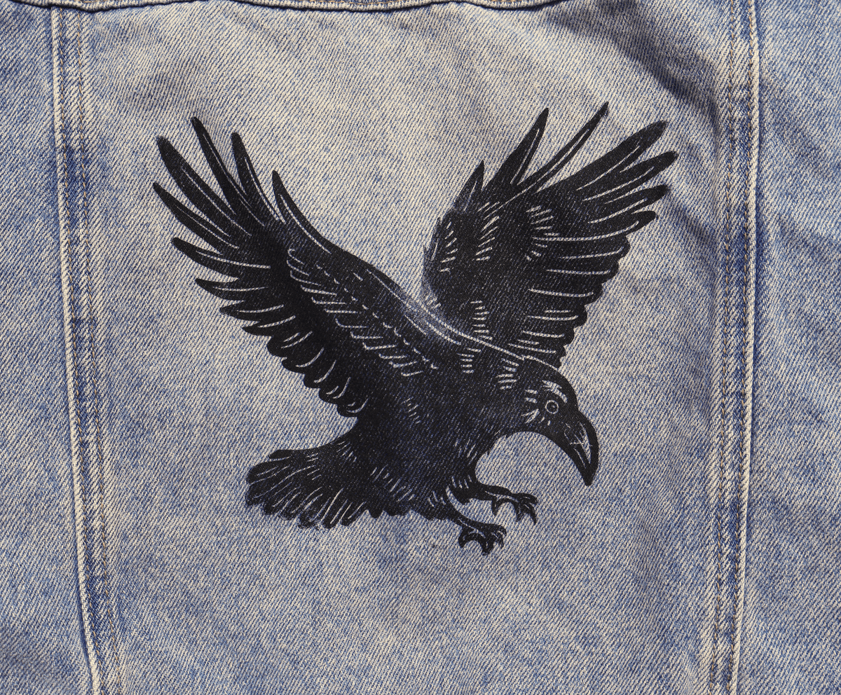 A close-up of light blue denim fabric featuring a large black embroidered raven with wings spread, stitched onto the back panel.