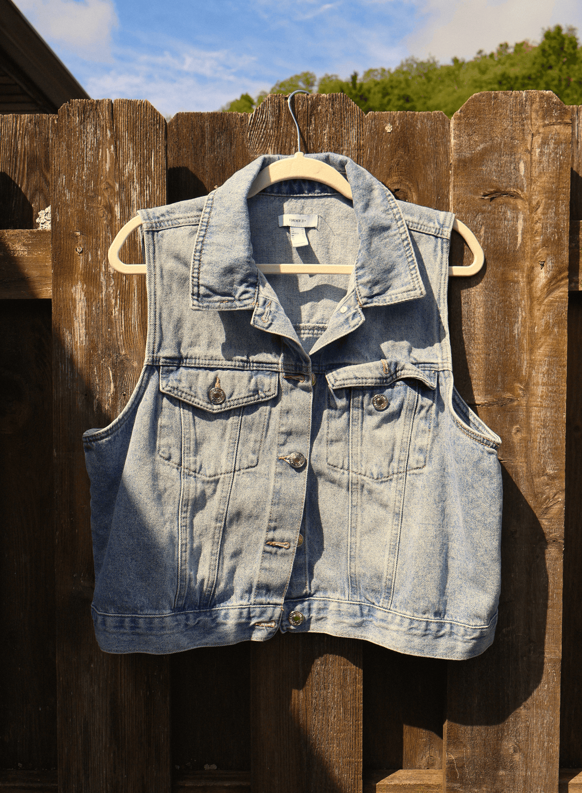 A light-wash denim vest hanging on a wooden hanger against a weathered brown wooden fence, with trees and blue sky visible in the background.