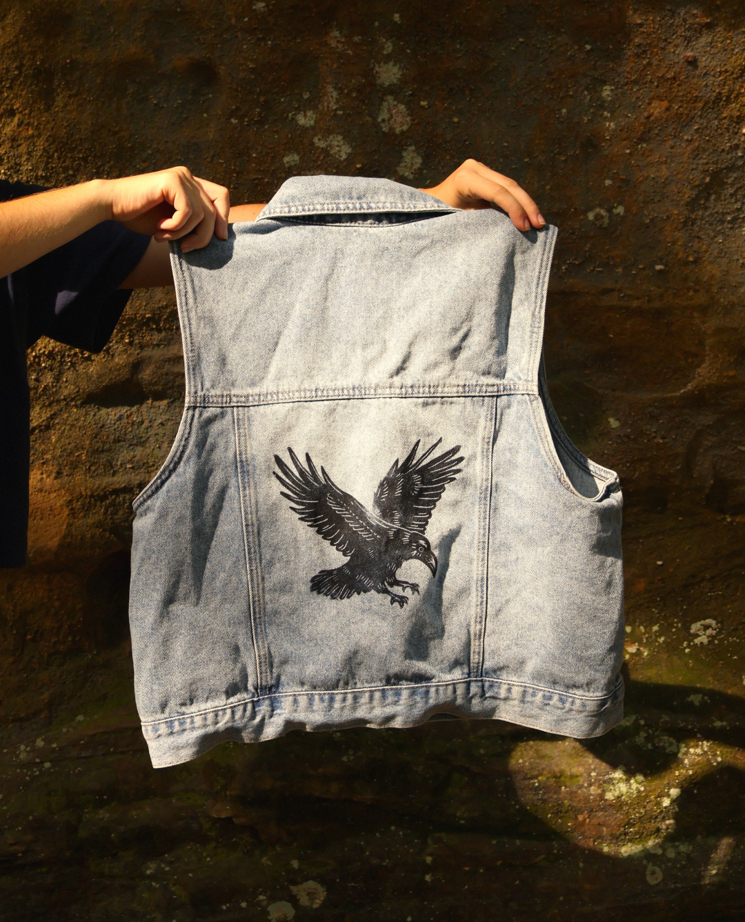 A light-wash denim vest is held up by two hands against a dark, textured stone wall. The back of the vest features a large black graphic print of a flying raven or crow.