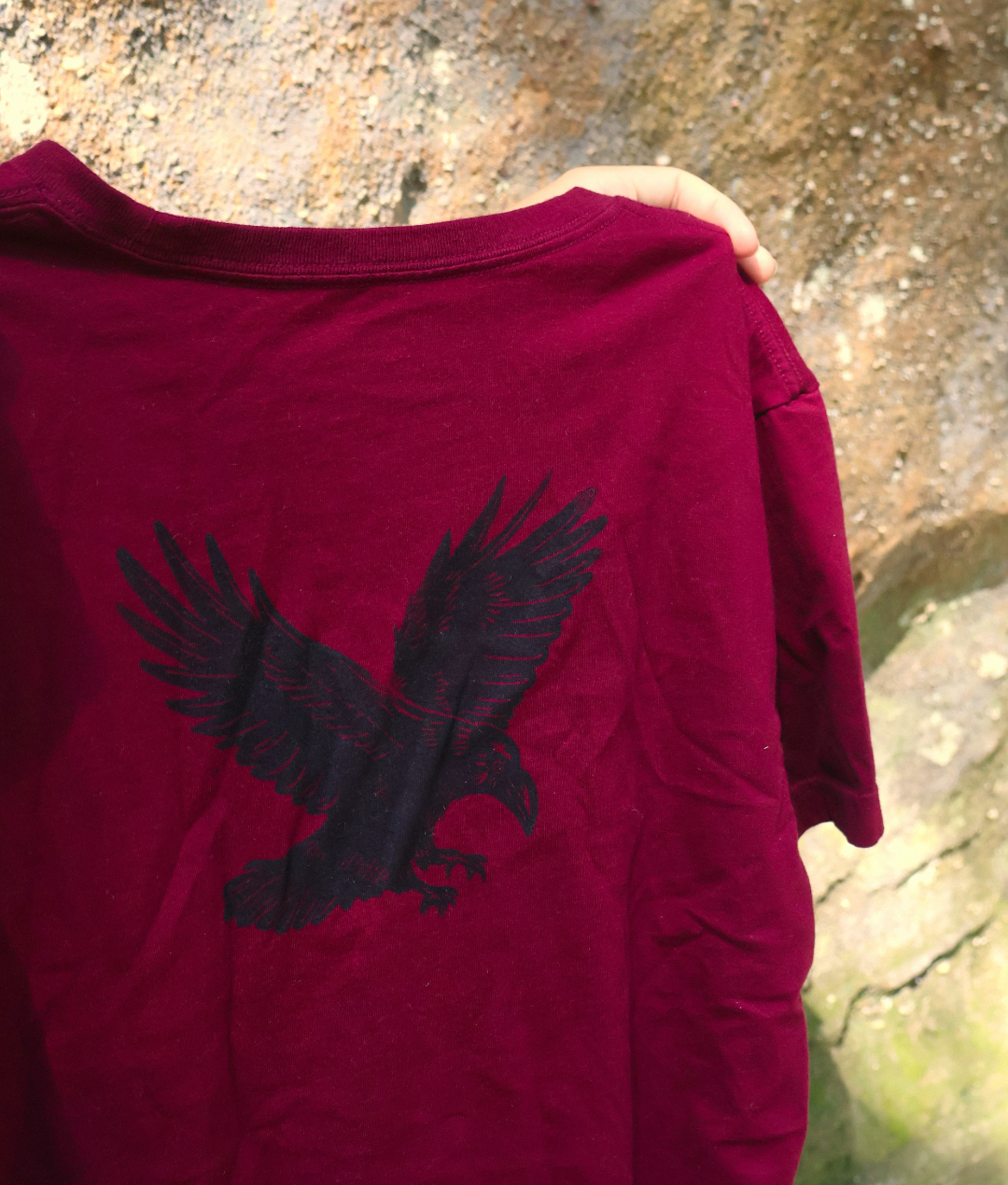 A close-up of a person holding a dark red t-shirt with a black eagle graphic on the back, set against a natural rocky background.