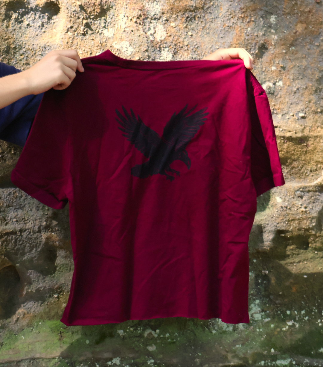 A close-up of a person holding a dark red t-shirt with a black eagle graphic on the back, set against a natural rocky background.