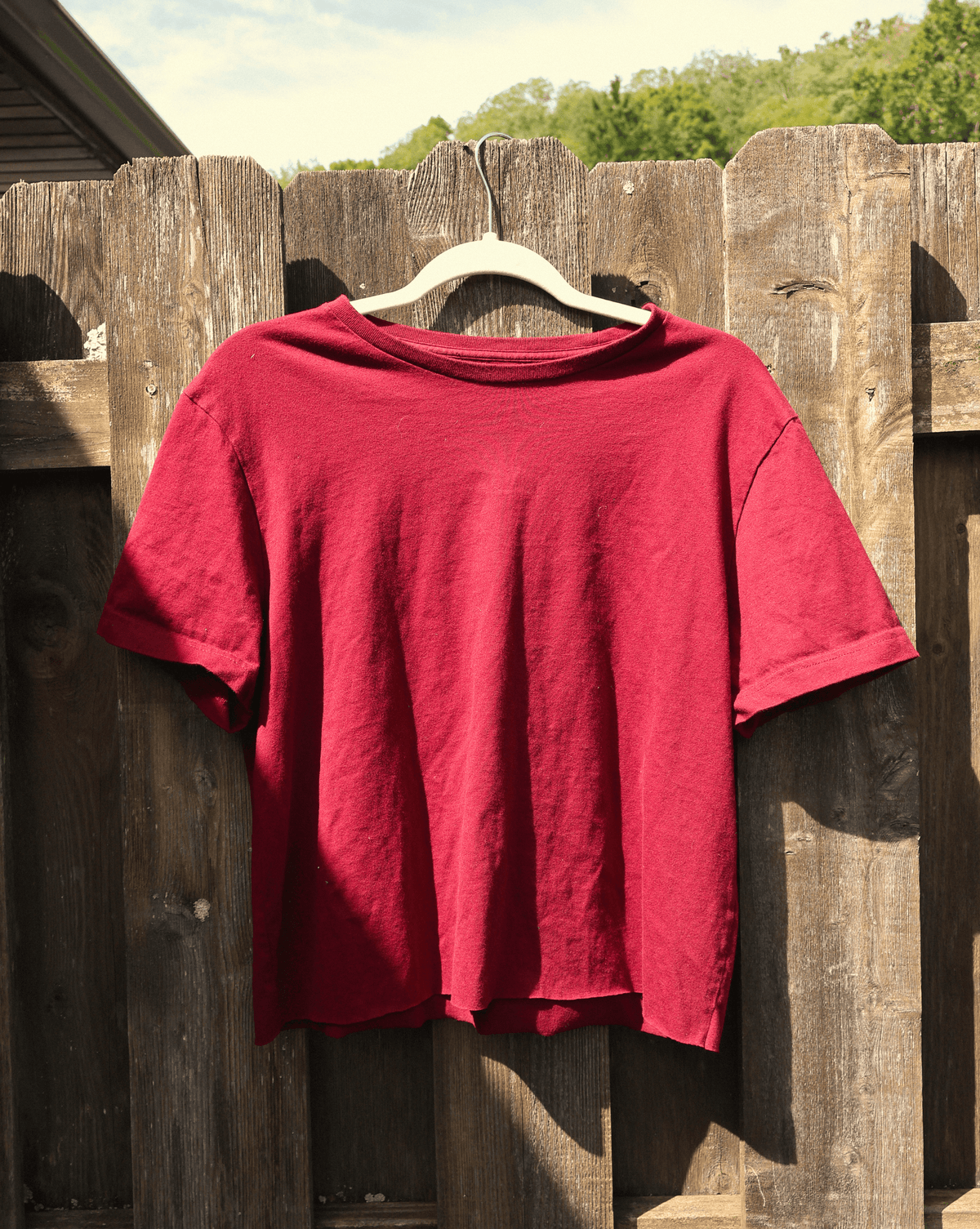 A red short-sleeve crewneck t-shirt hanging on a white plastic hanger against a weathered wooden fence outdoors.