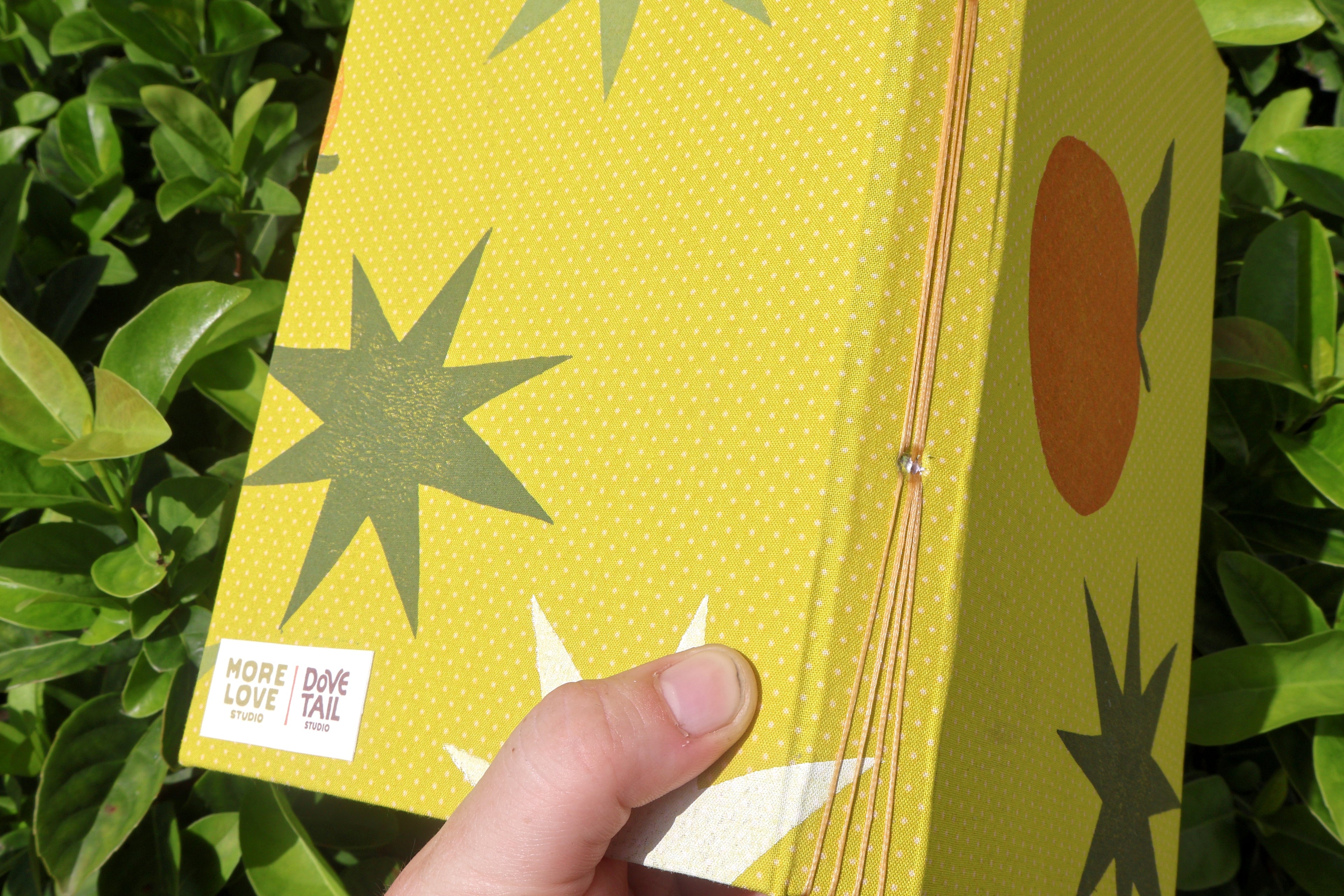 A hand holding a yellow patterned book or notebook outdoors in front of green leaves. The cover has a dotted yellow background with dark green starburst shapes, a large orange circle, and a white label reading 'MORE LOVE Dove tail'. Hand printed and bound self care eflections log