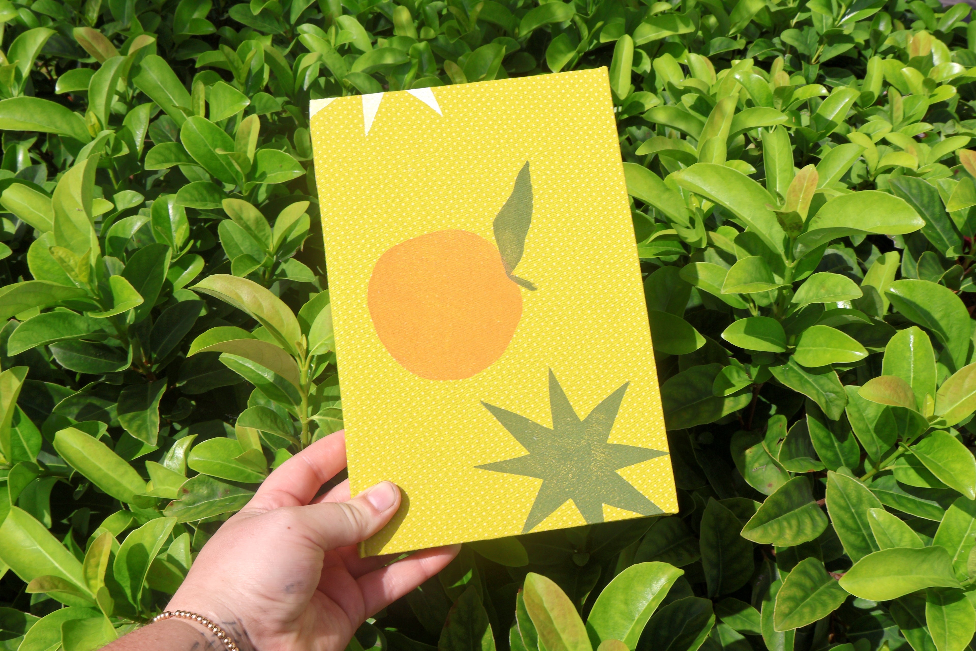 A hand holds a yellow rectangular card or notebook cover featuring a simple orange and green starburst illustration, set against a background of green leaves.