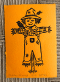 An orange art print featuring a hand-drawn black ink scarecrow with a bird on its shoulder, placed on a textured gray fabric background.