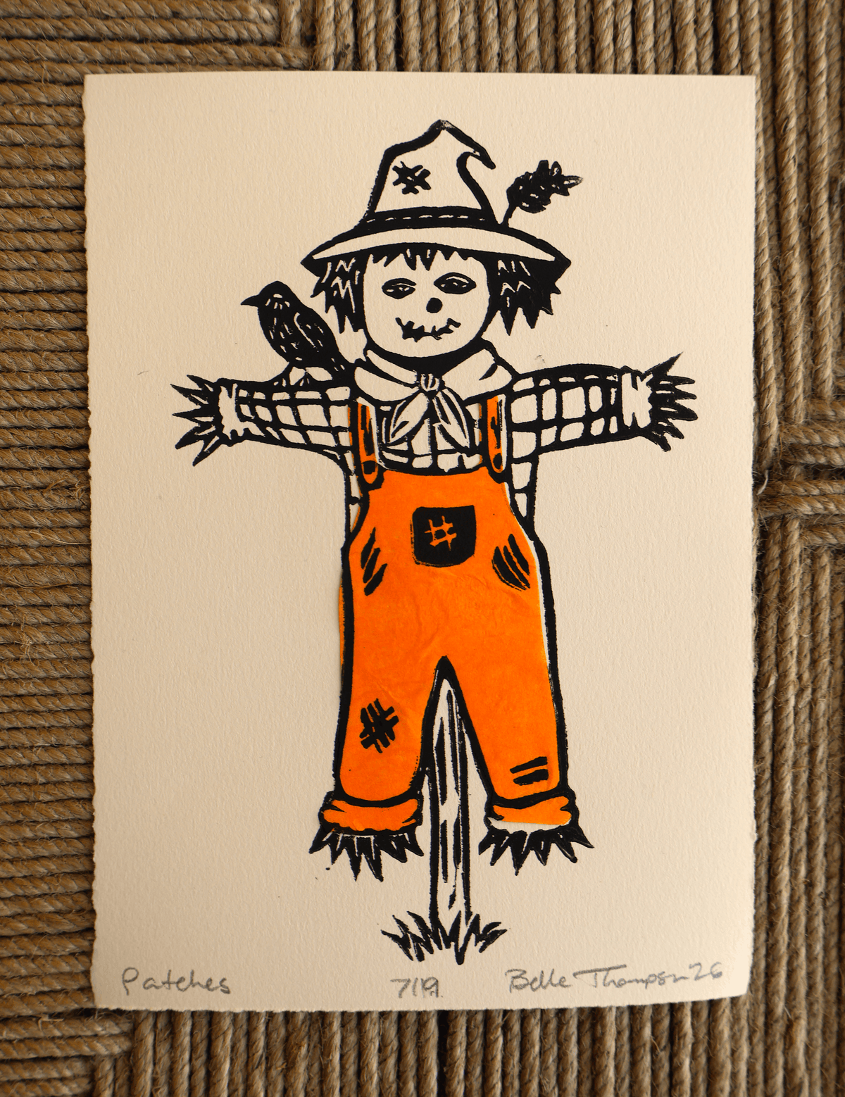 A handmade art print featuring a whimsical hand-drawn scarecrow on textured off-white paper, placed on a brown woven straw mat. The print is signed 'Patches 719 Belle Thompson 26' at the bottom.