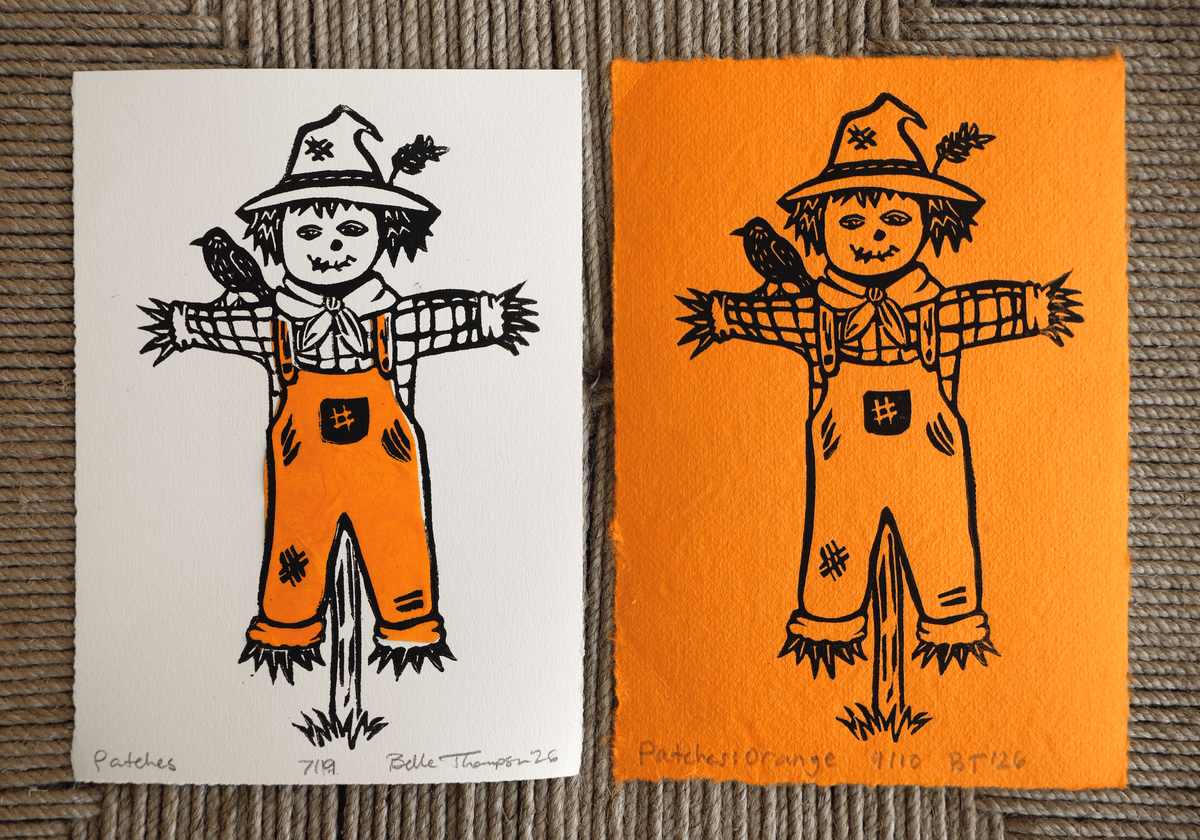 Two hand-signed and numbered art prints of a scarecrow with a bird on its shoulder, displayed side-by-side on a gray woven fabric background. The left print is on white paper and the right print is on orange paper.