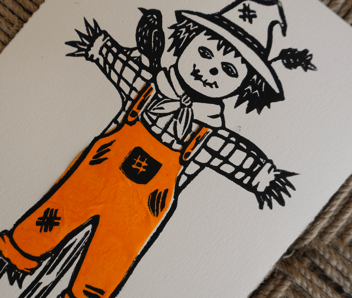 A close-up of a hand-painted scarecrow illustration on a white square canvas, featuring black ink lines and bright orange body paint, resting on a woven brown surface.