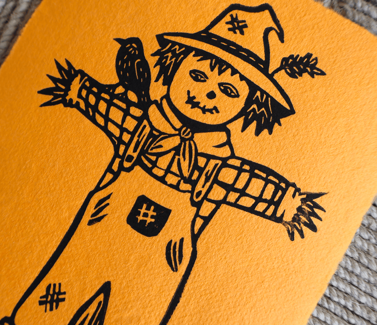 A close-up of a handmade orange card or art print featuring a black ink illustration of a scarecrow with a bird on its shoulder, set against a textured gray background.
