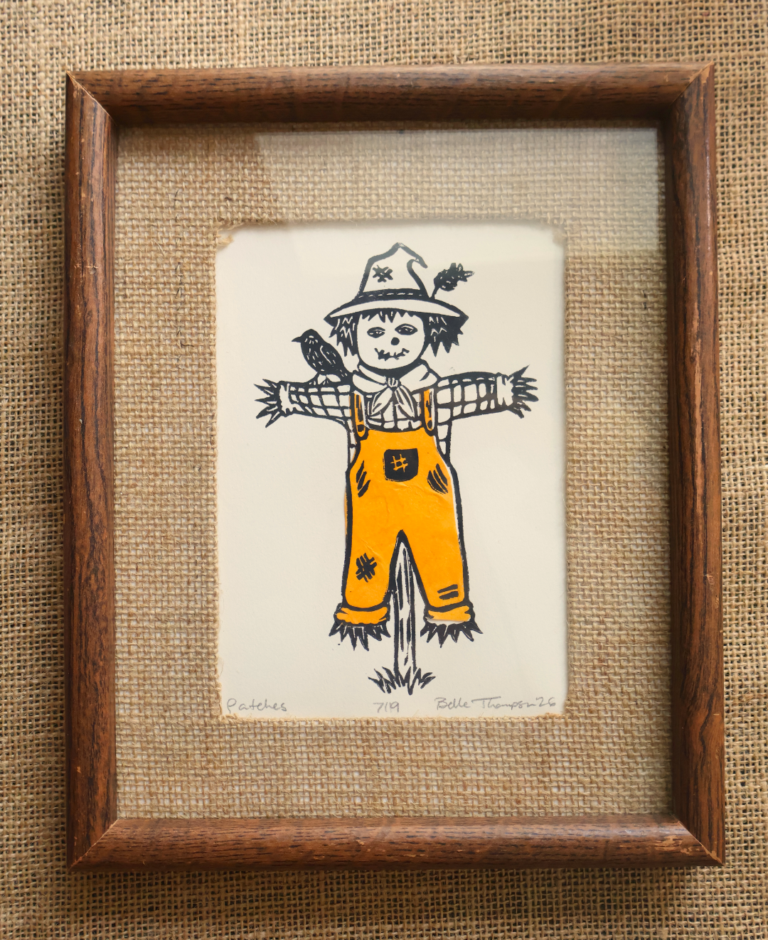 A handmade art print featuring a whimsical hand-drawn scarecrow on textured off-white paper, placed on a brown woven straw mat. The print is signed 'Patches'