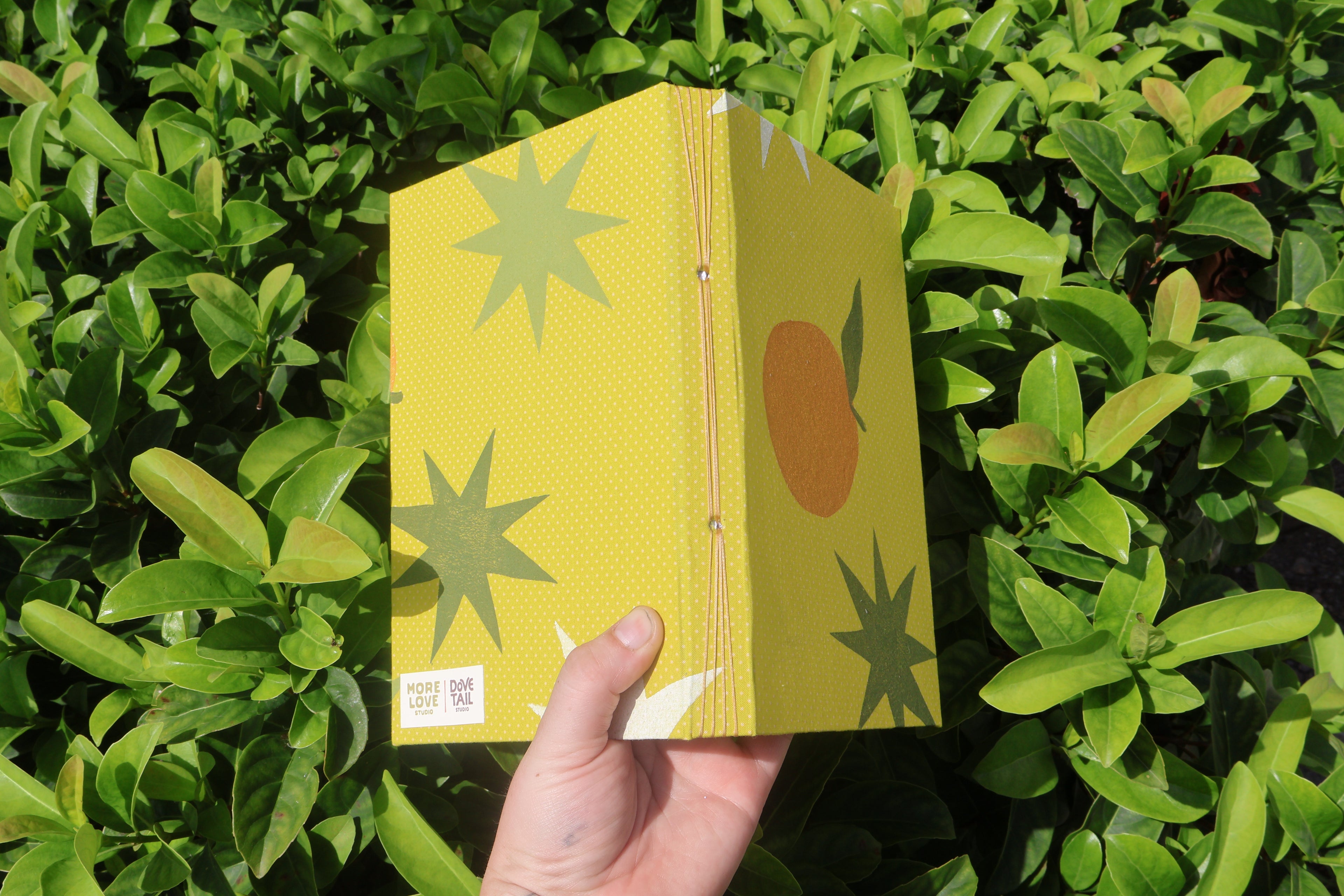 A hand holds a yellow patterned notebook or book cover in front of dense green foliage. The cover features a yellow background with a fine dot texture, olive green starburst shapes, a brown-orange circle with a green leaf, and a small white square label near the bottom left. hand printed and bound self care reflections log