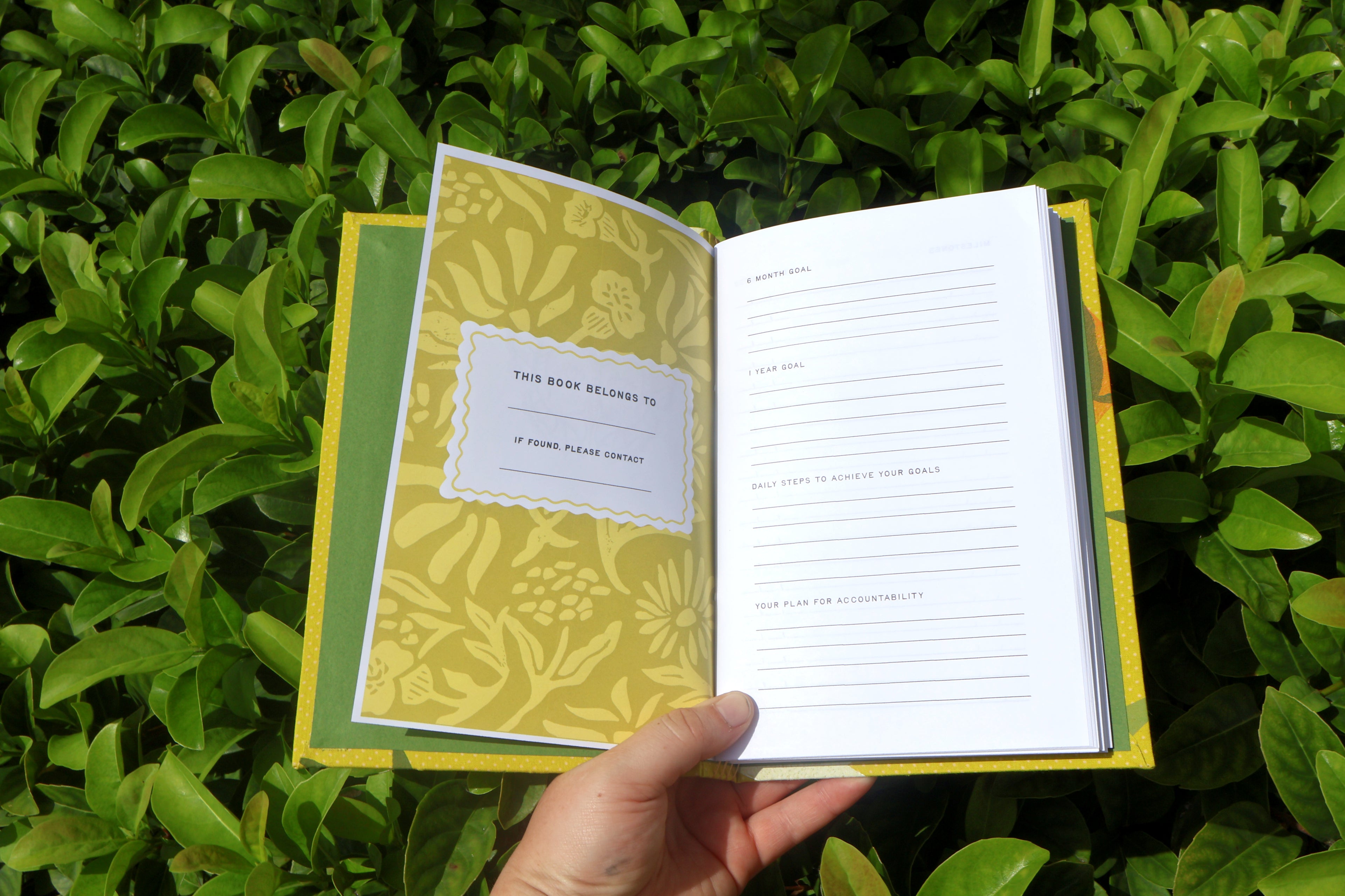 An open book or journal held by a hand, showing a yellow floral-patterned cover and white interior pages, photographed outdoors against dense green foliage. Hand printed and bound self care reflections log