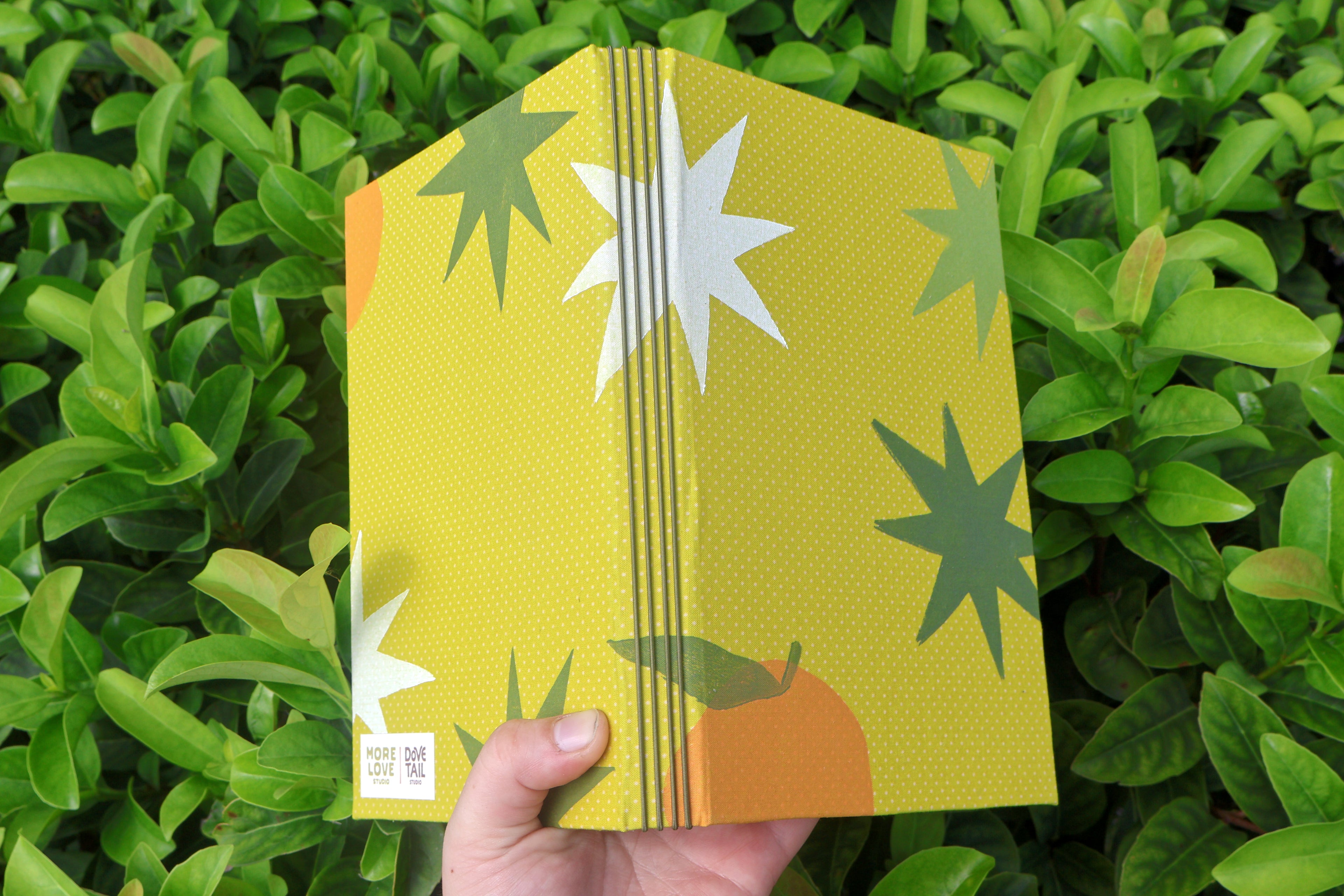 A hand holds the spine of yellow-patterned refillable bullet journals in front of dense green foliage. The journal has a white starburst, a dark green starburst, and a small orange shape. hand bound and linocut printed with more love studio