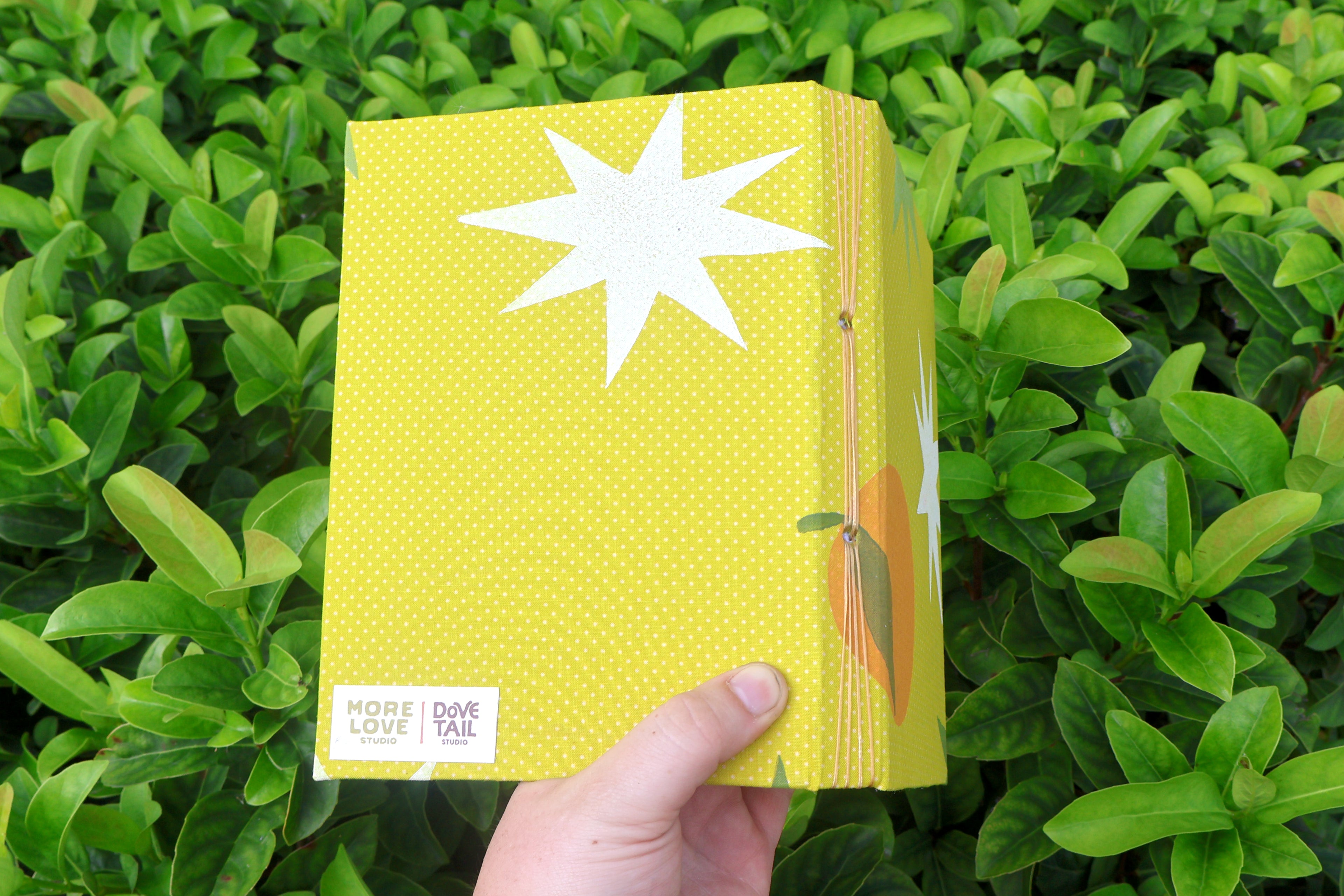 A hand holds an open, green, hand-bound and printed self-care reflection logs with white starburst shapes, photographed outdoors against dense green leaves. Hand bound and linocut printed with more love studio